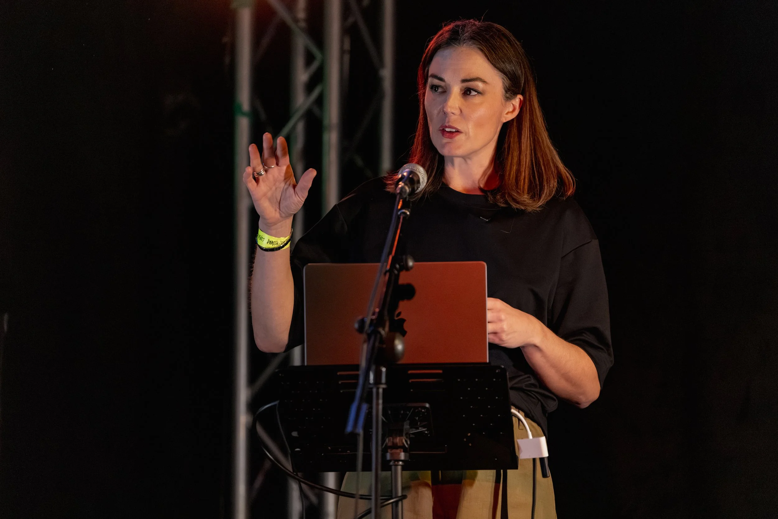 Katy Cowan speaking at a microphone during an event, with a laptop in front of her and a dark background.