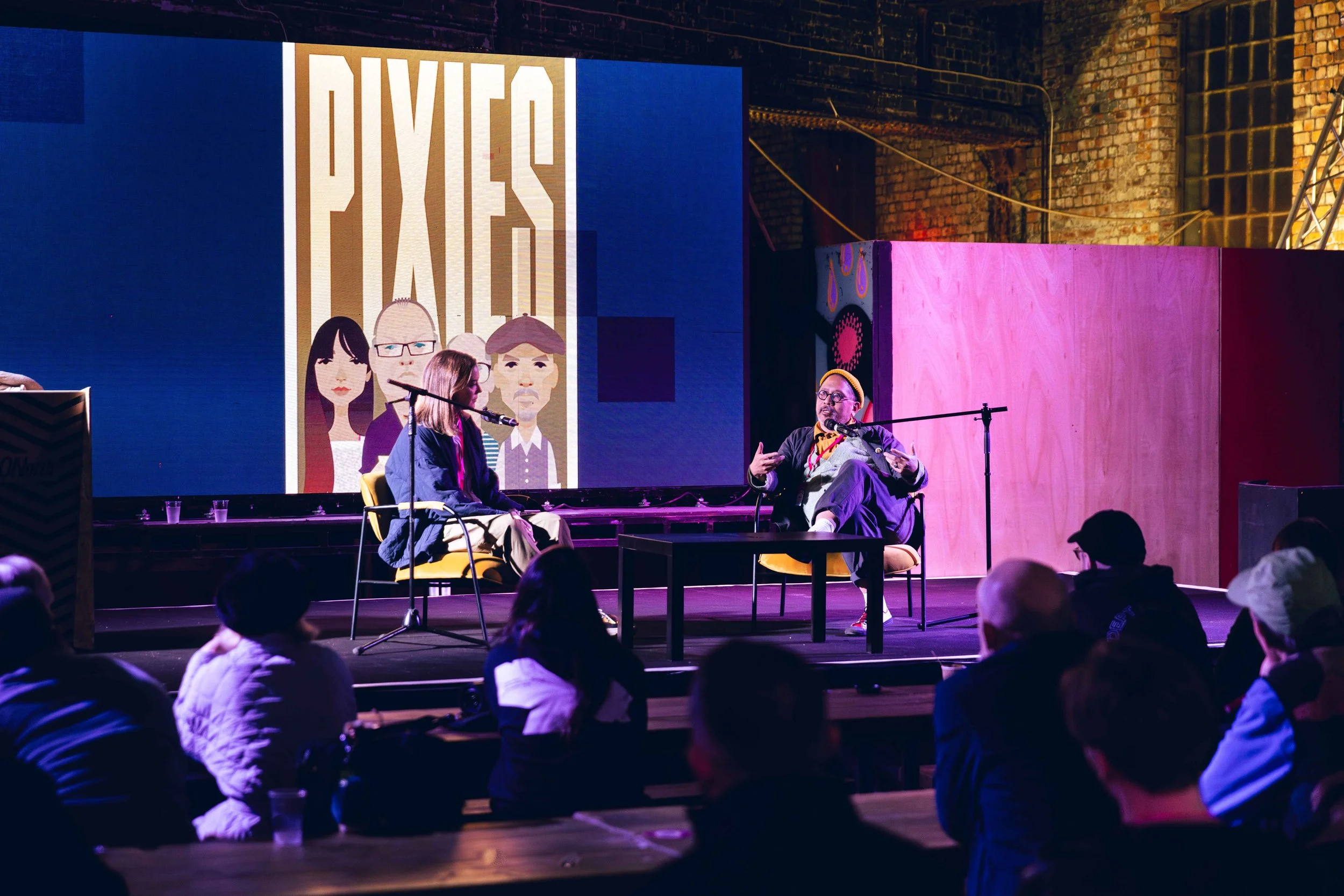 Katy Cowan and Stanley Chow are seated on stage in front of an audience, engaged in a discussion or interview, with a large screen displaying animated graphics and the word 'PULSE' in the background.