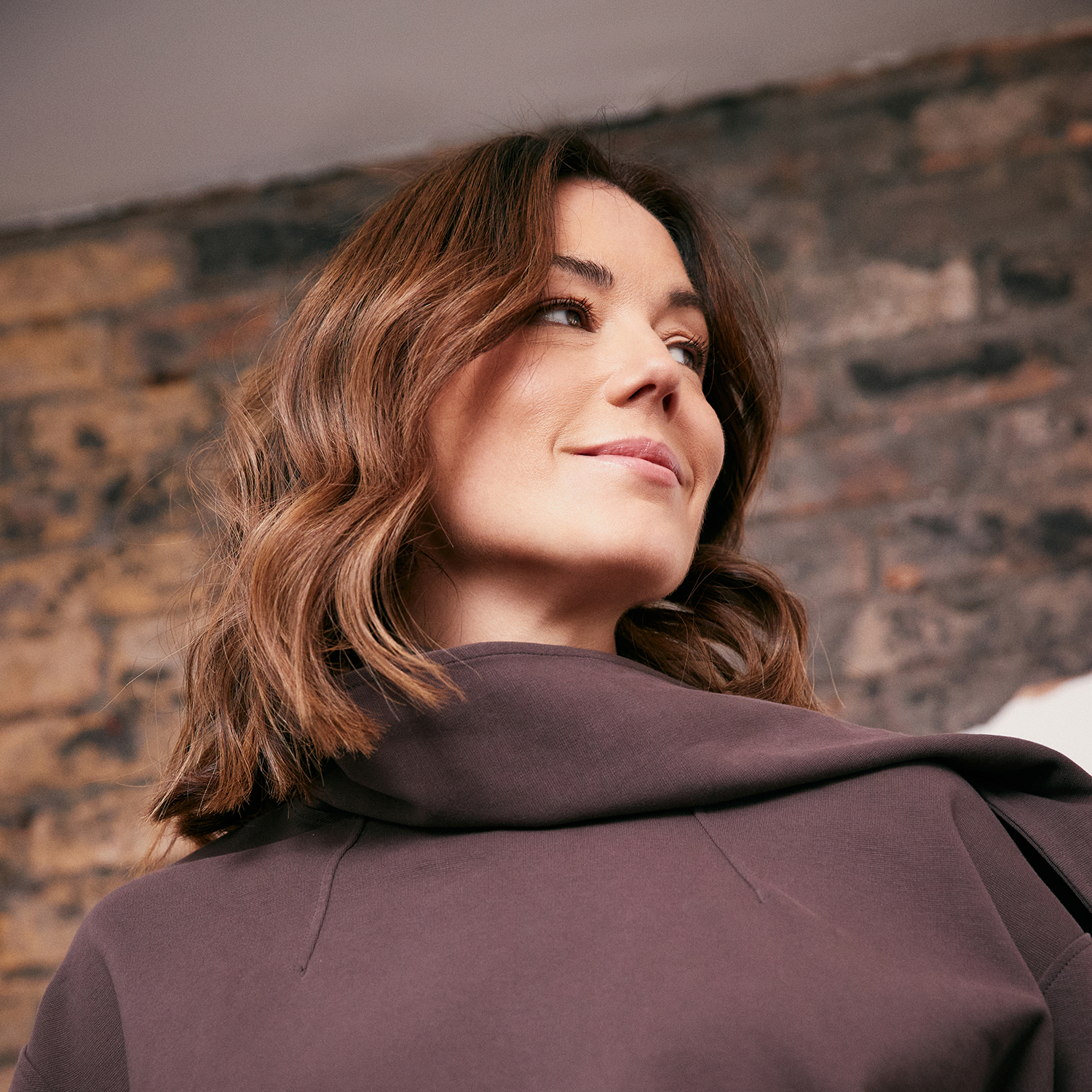 Katy Cowan, a woman with wavy brown hair, smiling slightly, turned over her shoulder, wearing a dark brown top, with a brick wall in the background.