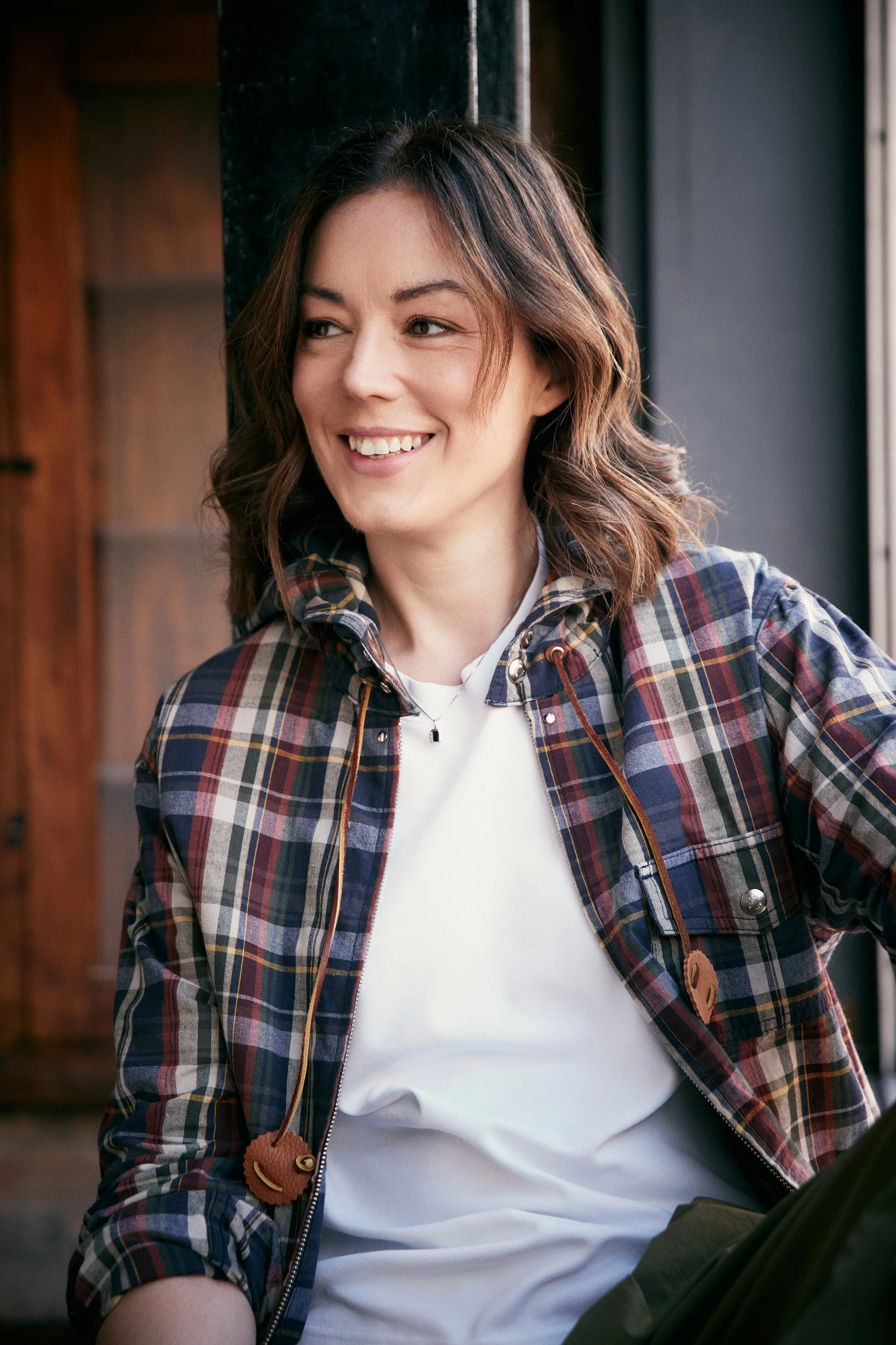 Katy Cowan, with shoulder-length brown hair, smiling while sitting indoors near a window, wearing a plaid shirt over a white T-shirt.
