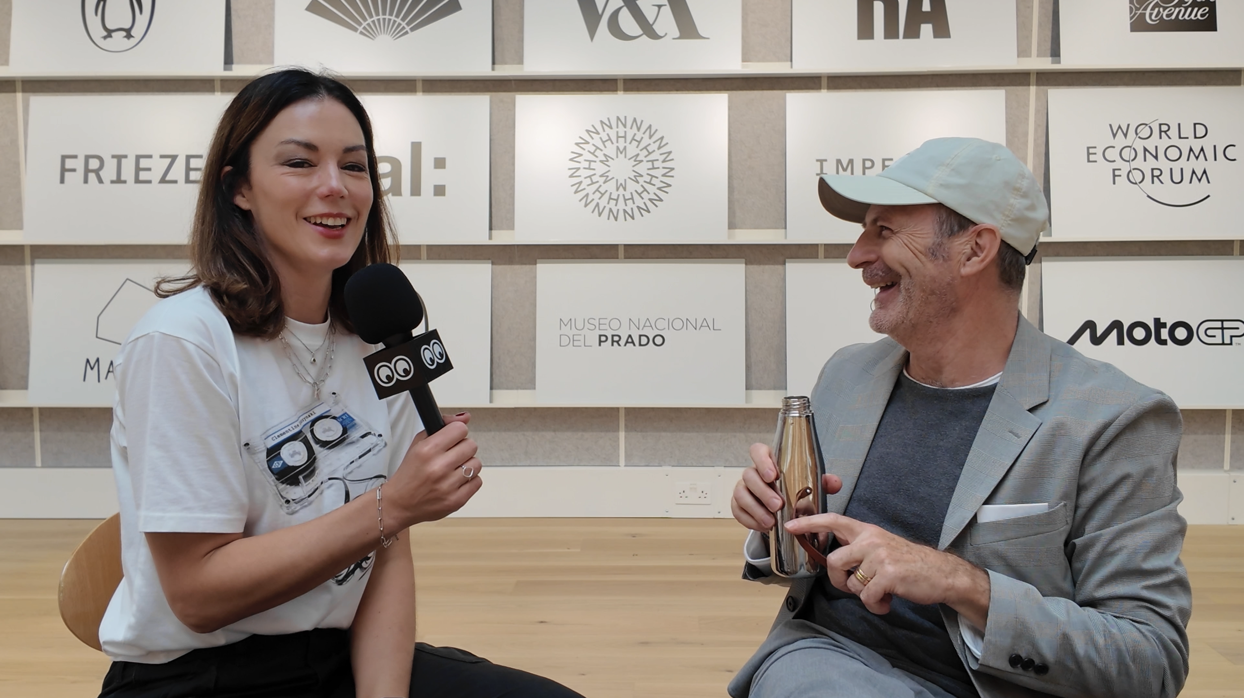 Katy Cowan is holding a microphone, sitting and smiling during an interview or conversation with Angus Hyland of Pentagram. They are in front of a wall with logos and text related to the World Economic Forum and Museo Nacional del Prado.