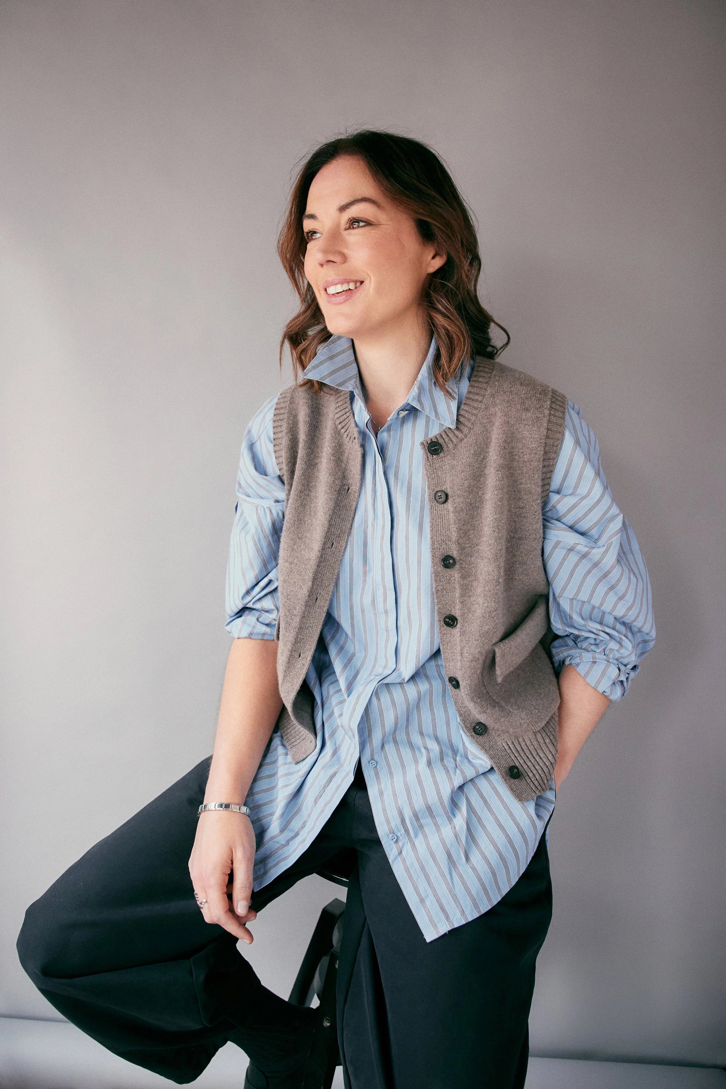 Katy Cowan, a woman with shoulder-length brown hair in a loose wave, wearing a blue striped shirt layered under a grey sleeveless cardigan, sitting on a black stool against a plain grey background, smiling softly and looking to her left.