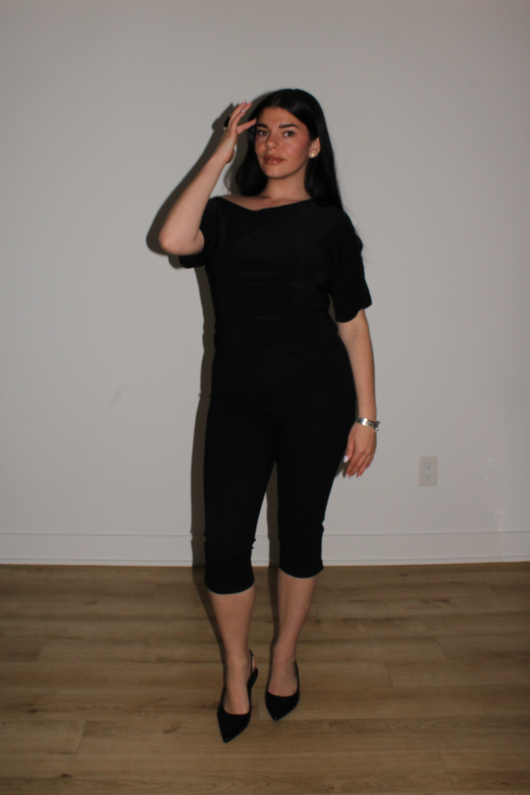 A woman with long black hair, wearing a black dress and black high heels, standing against a plain wall. She has one hand on her forehead and the other relaxed by her side. The floor is wood, and there is an electrical outlet near her feet.