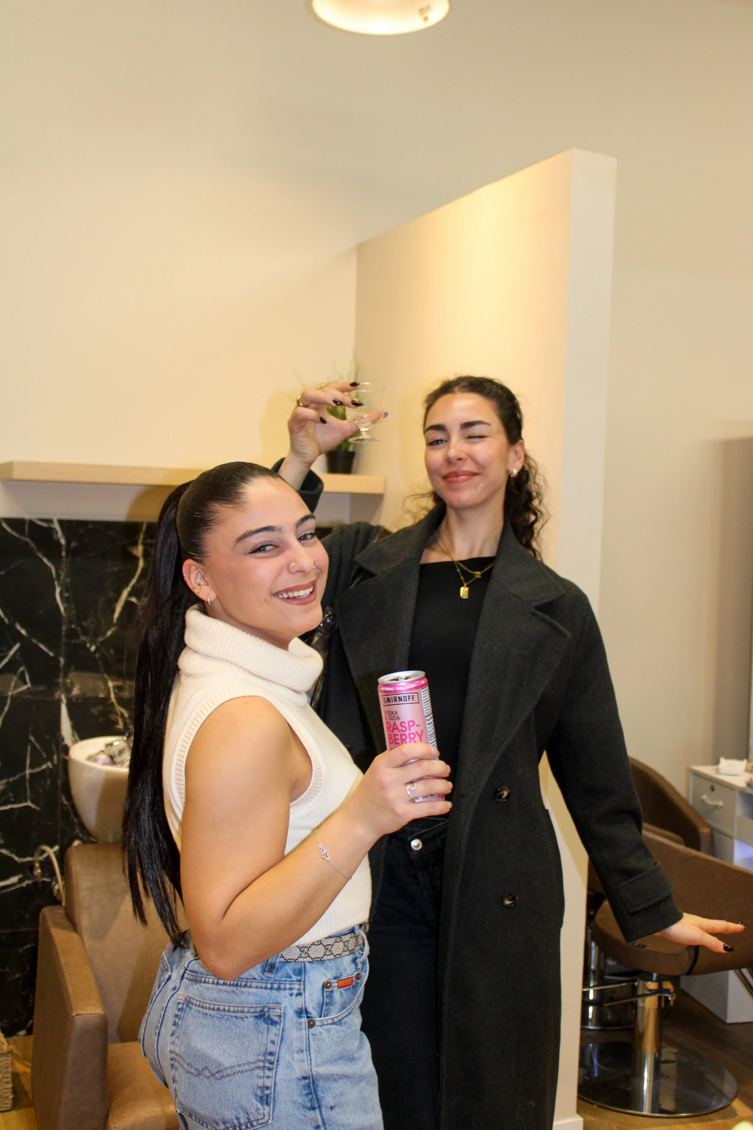 Two women celebrating with drinks in an indoor setting, one holding a can of Smirnoff Raspberry Fusion with a hand on her waist, smiling, and the other raising a cocktail glass, blushing, with a background of black marble and beige walls.