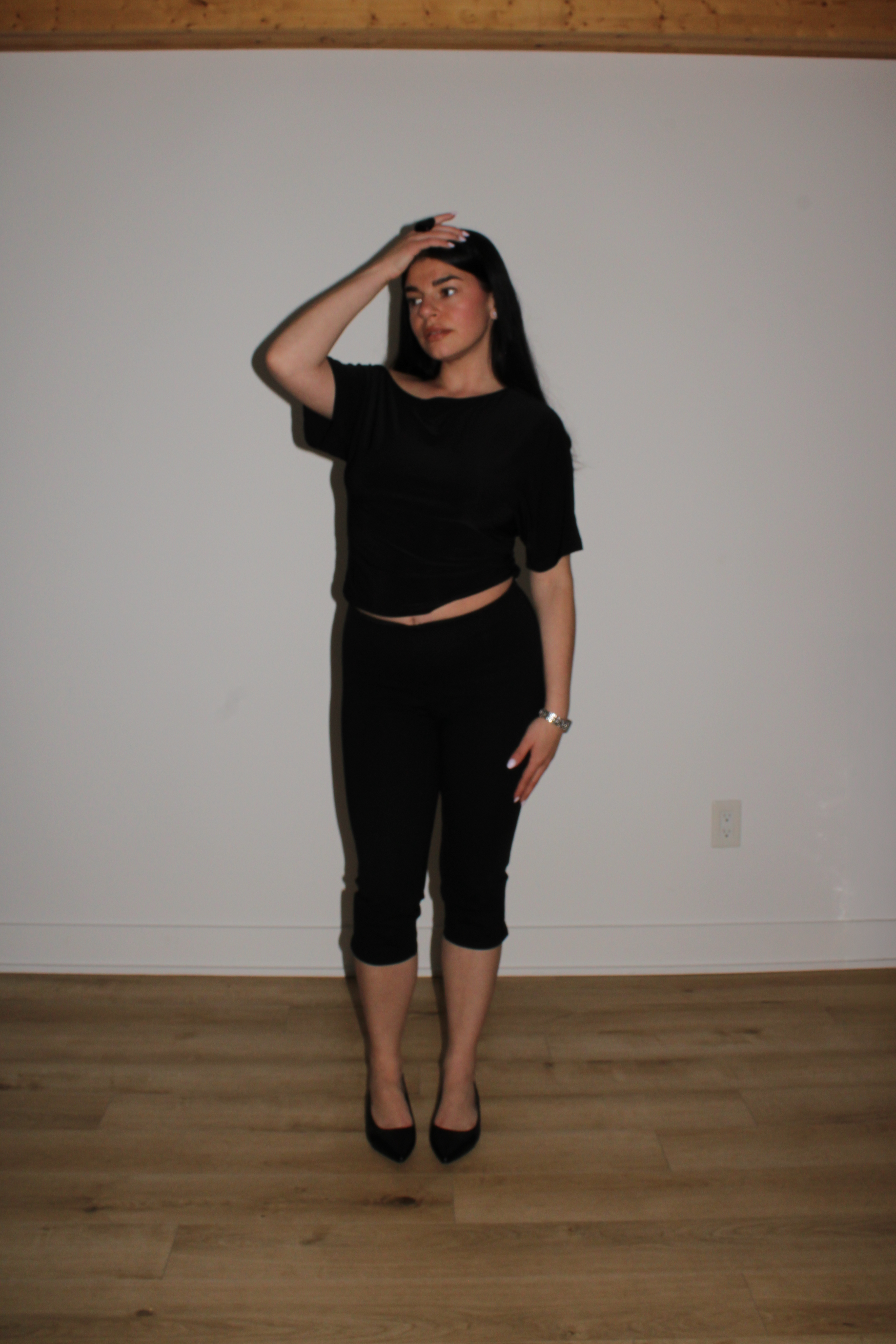 A woman with long black hair, dressed in black clothing, standing against a plain white wall. She is touching her forehead with her right hand and looking slightly to her left.