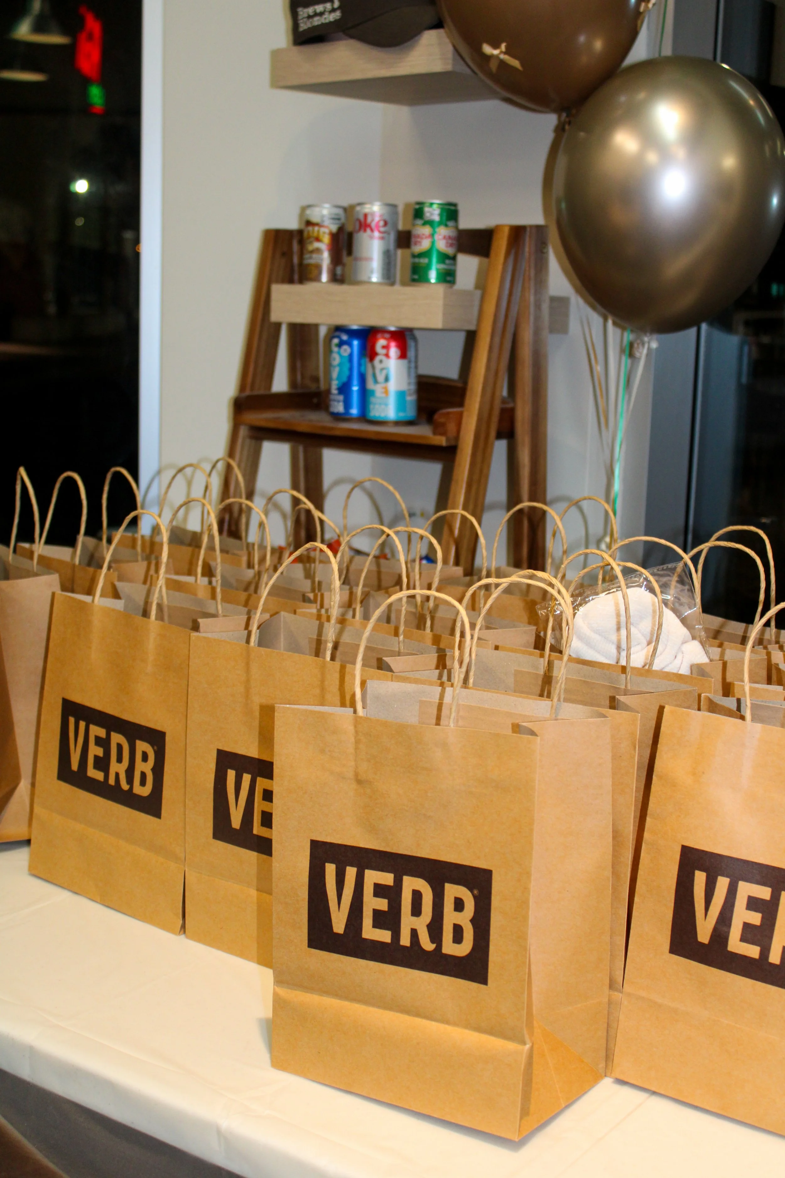 Brown paper bags with black and white VERB logo on a white table, balloons in the background, and canned beverages on a wooden shelf.