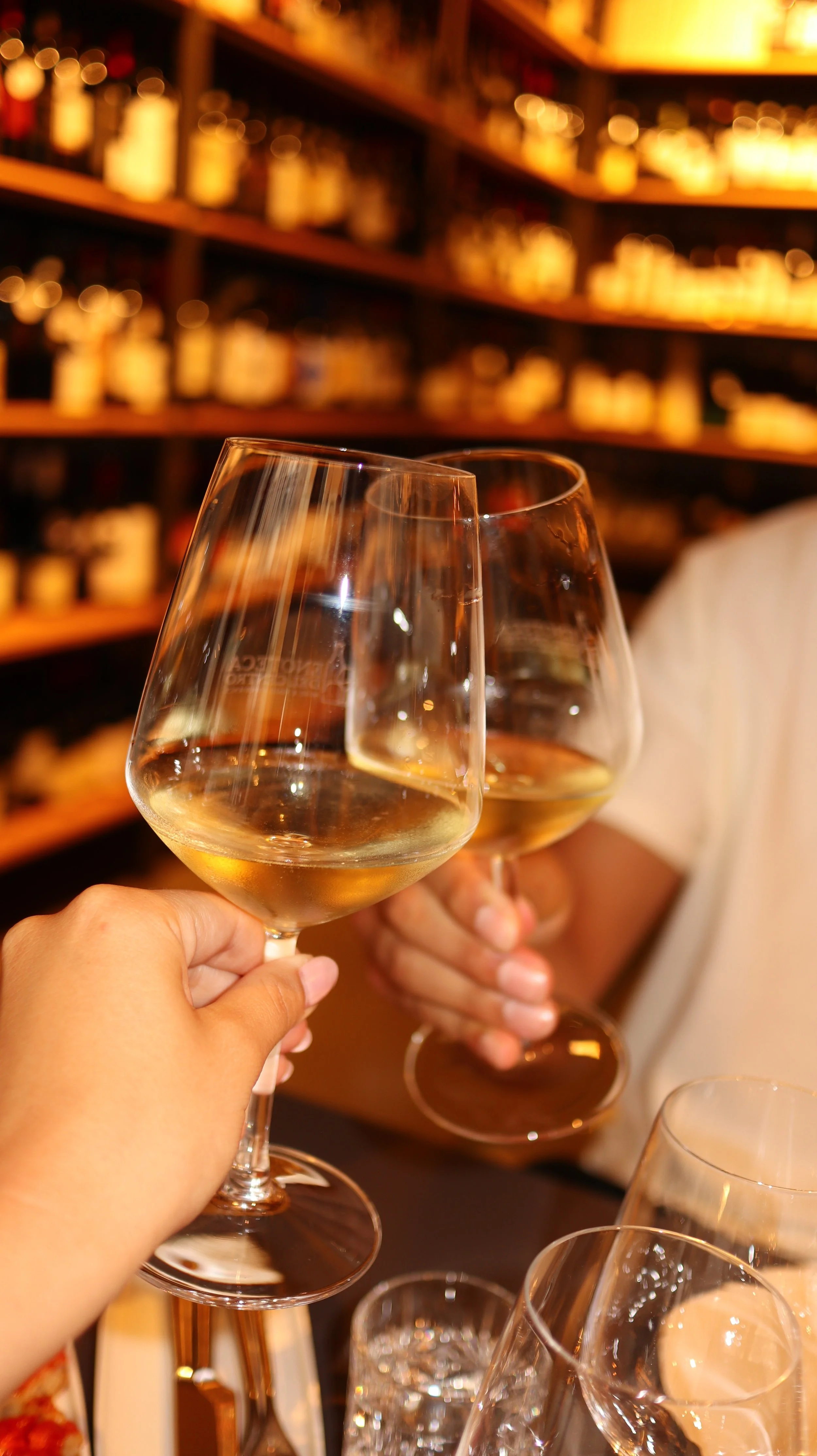 Two people clinking glasses of white wine in a bar or wine shop, with shelves filled with bottles in the background.