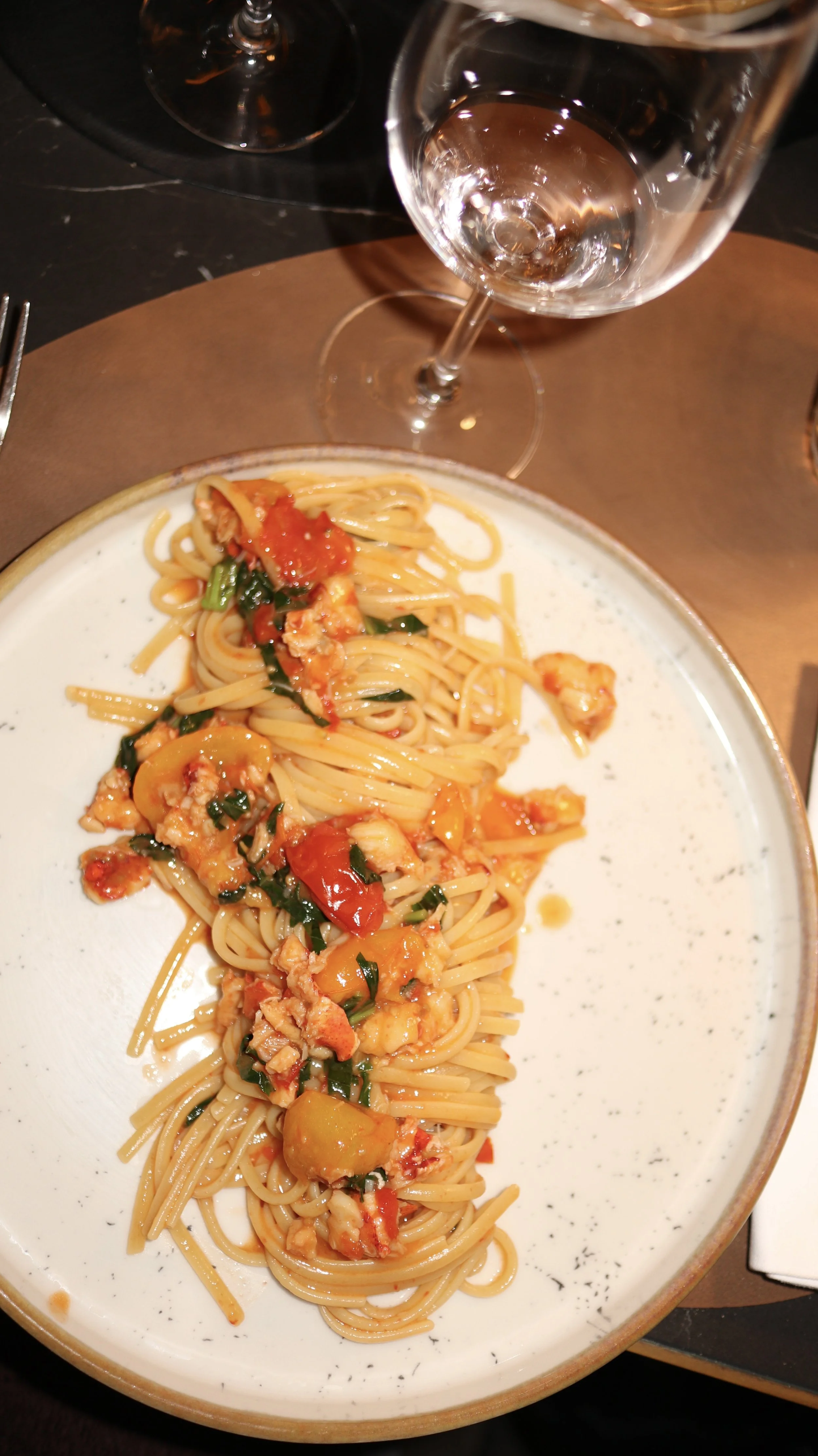 A plate of spaghetti with tomato sauce, vegetables, and herbs, next to two empty wine glasses on a dark table.