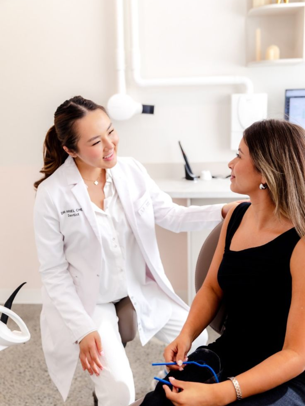 A female dentist in a white coat smiling and talking to a female patient in a dental office.