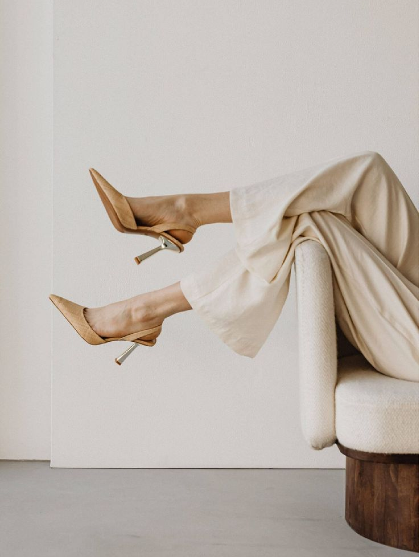 A person is sitting on a beige sofa with their legs crossed at the ankles and feet up, wearing beige high heels and a cream-colored dress. The photo is taken indoors with a plain light-colored wall in the background.