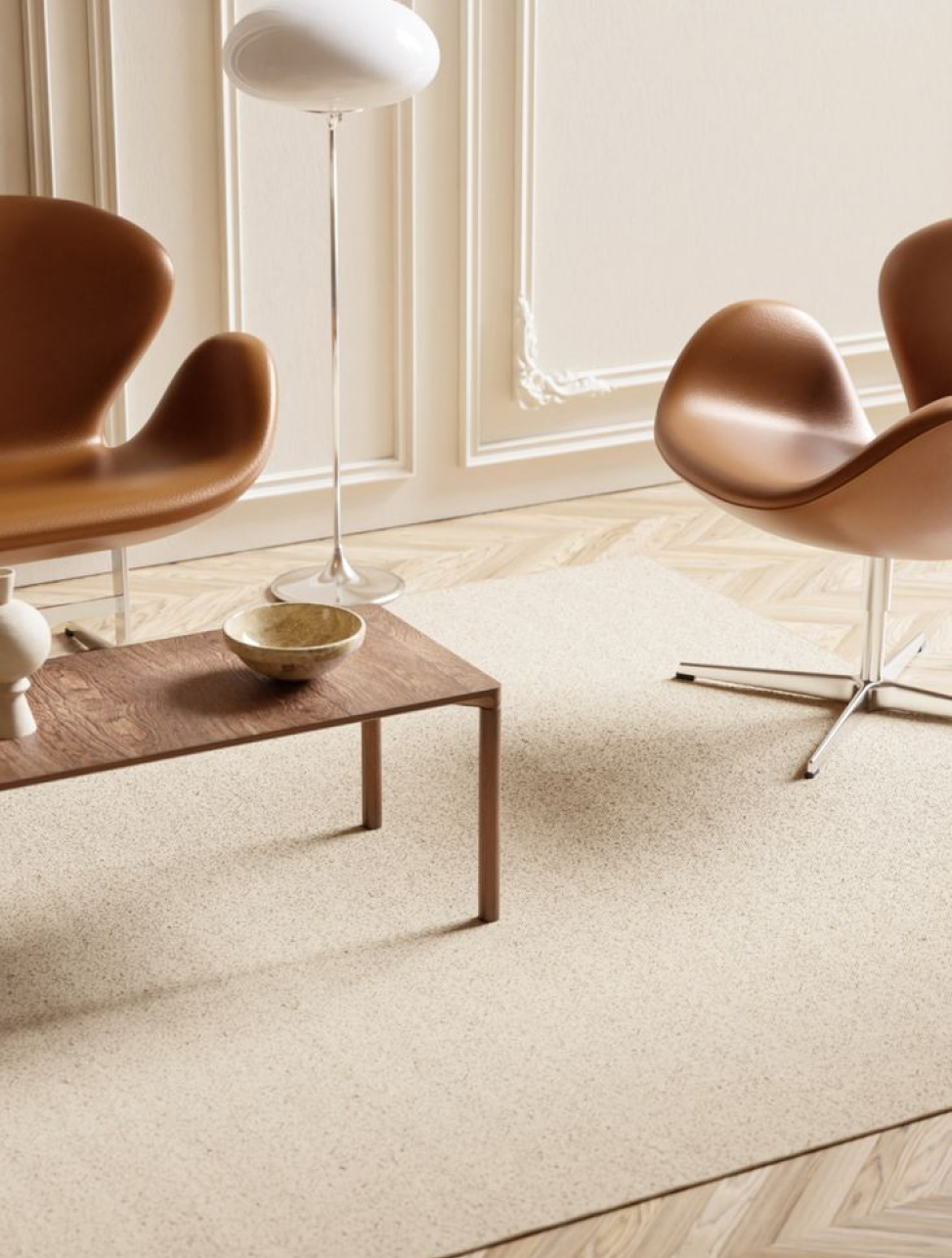 Interior living room with two brown swivel chairs, a wooden coffee table with a bowl, and a white floor lamp, beige carpet, and cream walls with decorative molding.