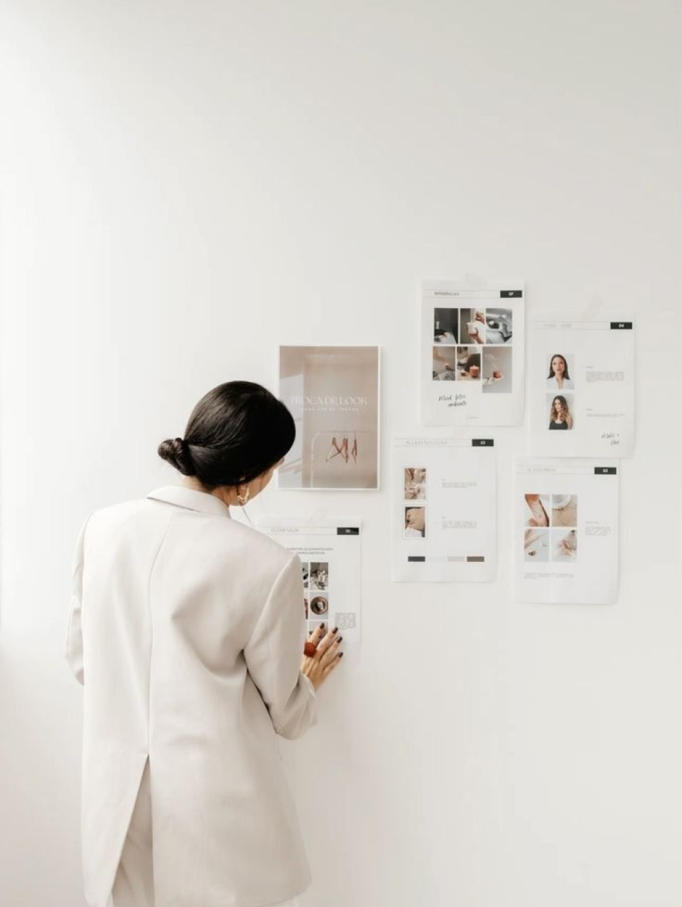 Woman in white blazer looking at a white wall with various printed documents and photos taped on it.