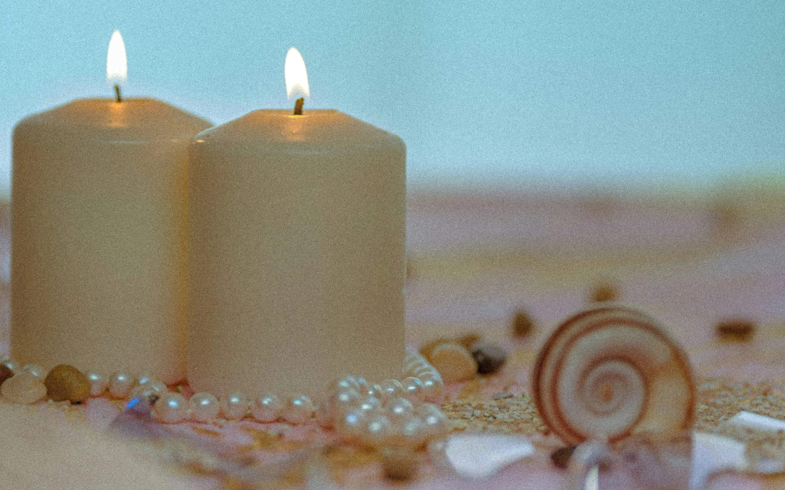Two beige candles with lit small flames, pearl jewelry, and assorted seashells on a pink sand surface with a blue water background.