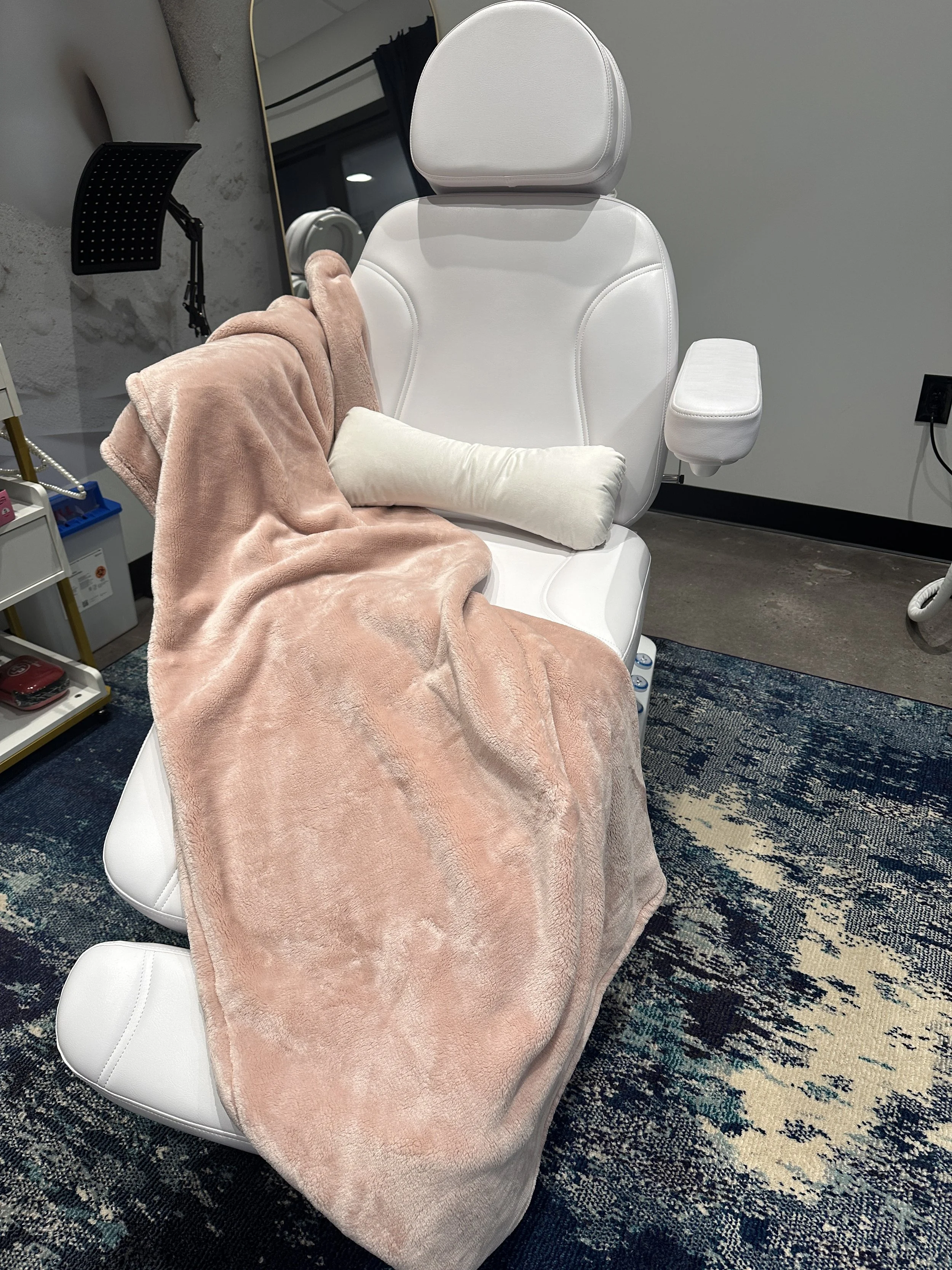 At Pearl Cove Aesthetics in Alpharetta, a white facial spa or medical chair with a pillow on the seat, draped with a pink fluffy blanket, in a room with a blue patterned rug, a mirror, and various items nearby.