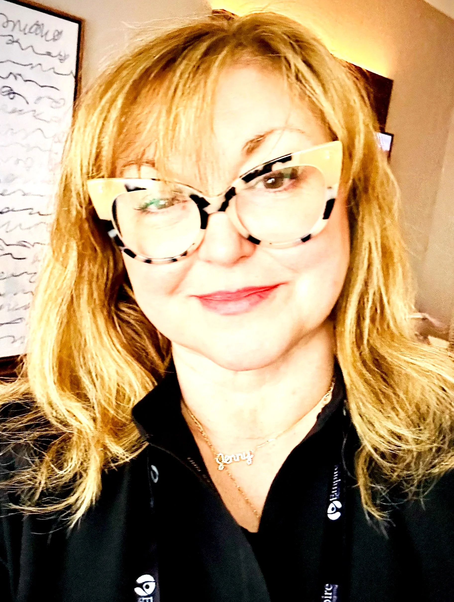 Close up of blond haired woman with tortoise shell glasses wearing a black scrub top with framed art and an edge of a tv frame in the background.