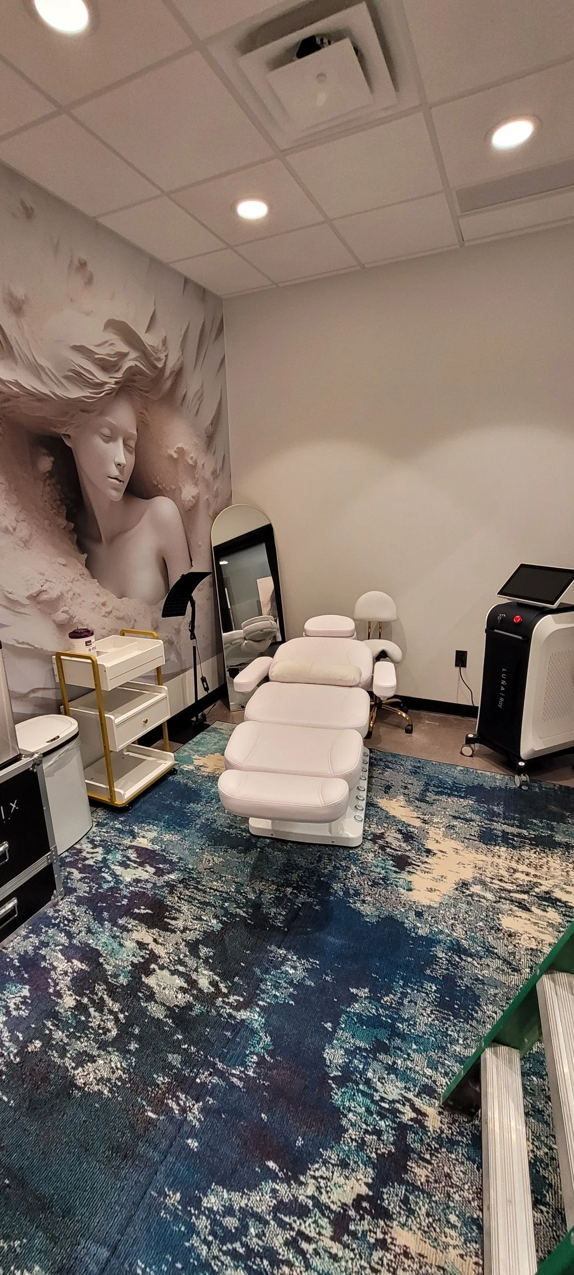 Spa treatment room with white facial chair, laser machine, mirror, storage tray, blue and white abstract rug with 3D mural of woman