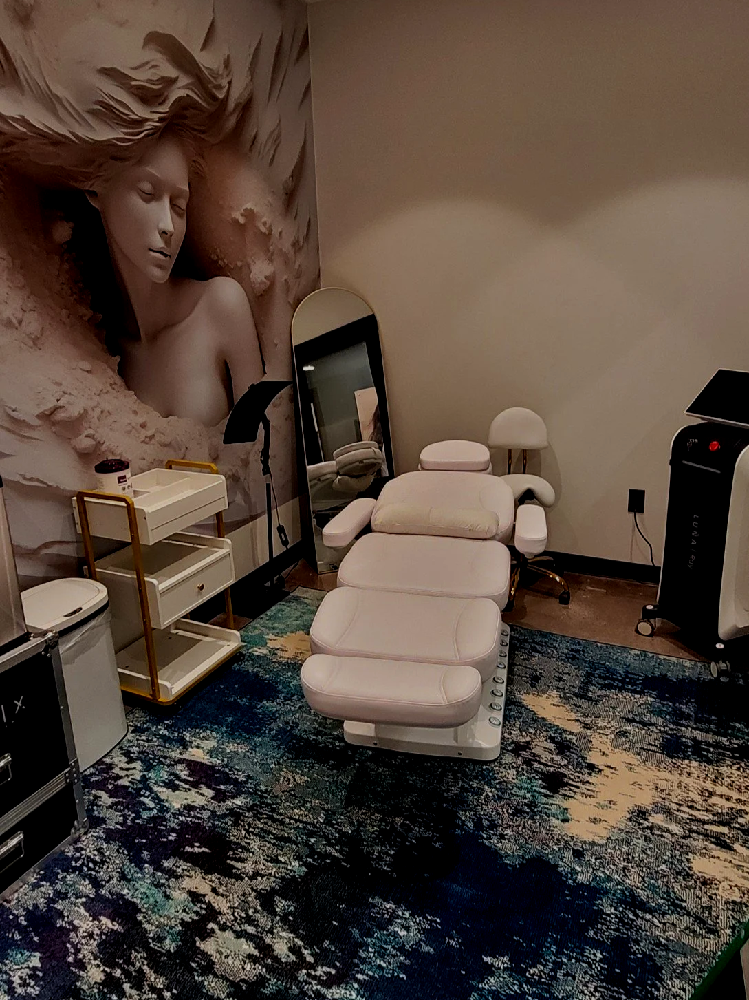 A massage chair in a room with a large mural of a woman with closed eyes and flowing hair, a mirror, a white trolley, a small white stool, a black and white machine, and a colorful rug.