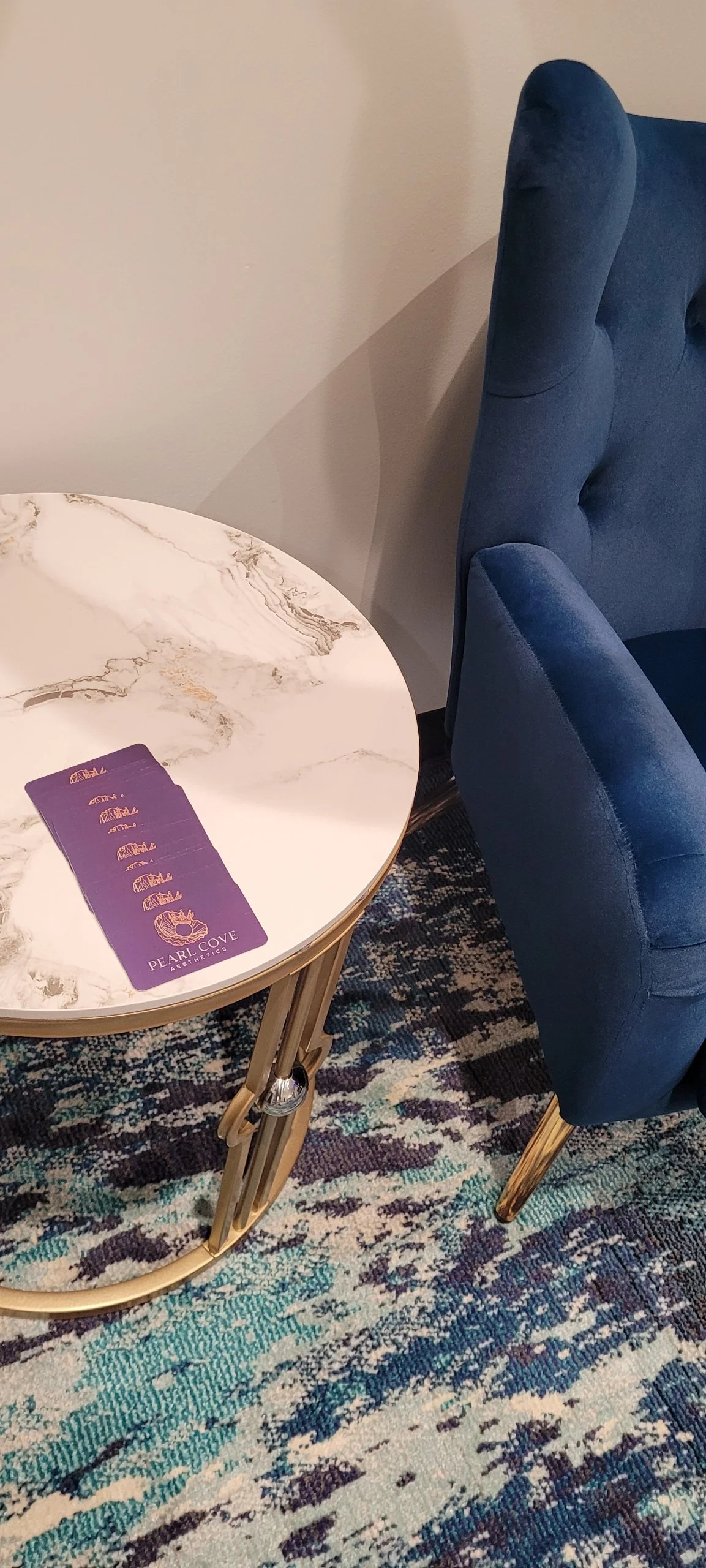 A marble-top table with purple business cards with reading Pearl Cove Aesthetics on it, next to a plush blue armchair, on a patterned carpet.