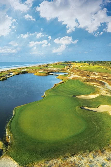 A scenic golf course with green fairways, sand traps, and a water hazard, overlooking the ocean under a partly cloudy sky.