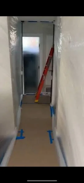 Narrow hallway under construction with a red ladder leaning against a mirror at the end, plastic sheeting covering the walls, and blue tape on the floor.
