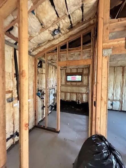 Interior of a house under construction with exposed wooden framing and insulation, unfinished walls, and a black trash bag on the floor.