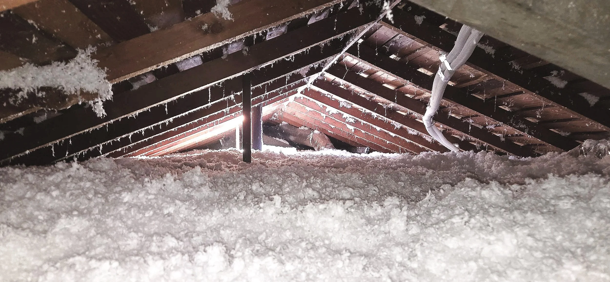 Attic with snow and ice buildup on the roof, showing exposed roof beams and a pipe, with sunlight shining through a small gap.
