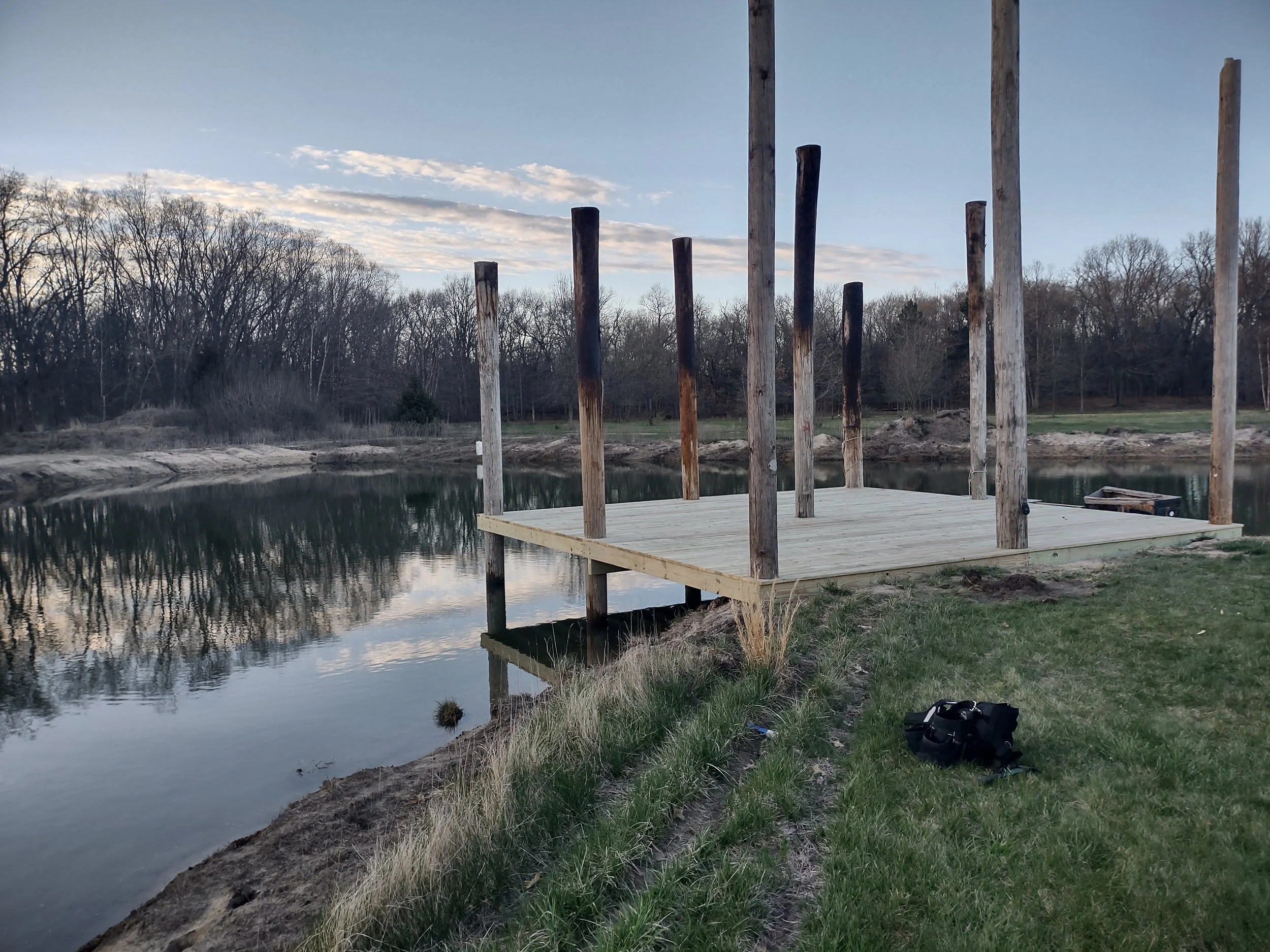 Unfinished wooden dock extending over a calm body of water with standing poles, surrounded by grassy land and leafless trees under a partly cloudy sky.