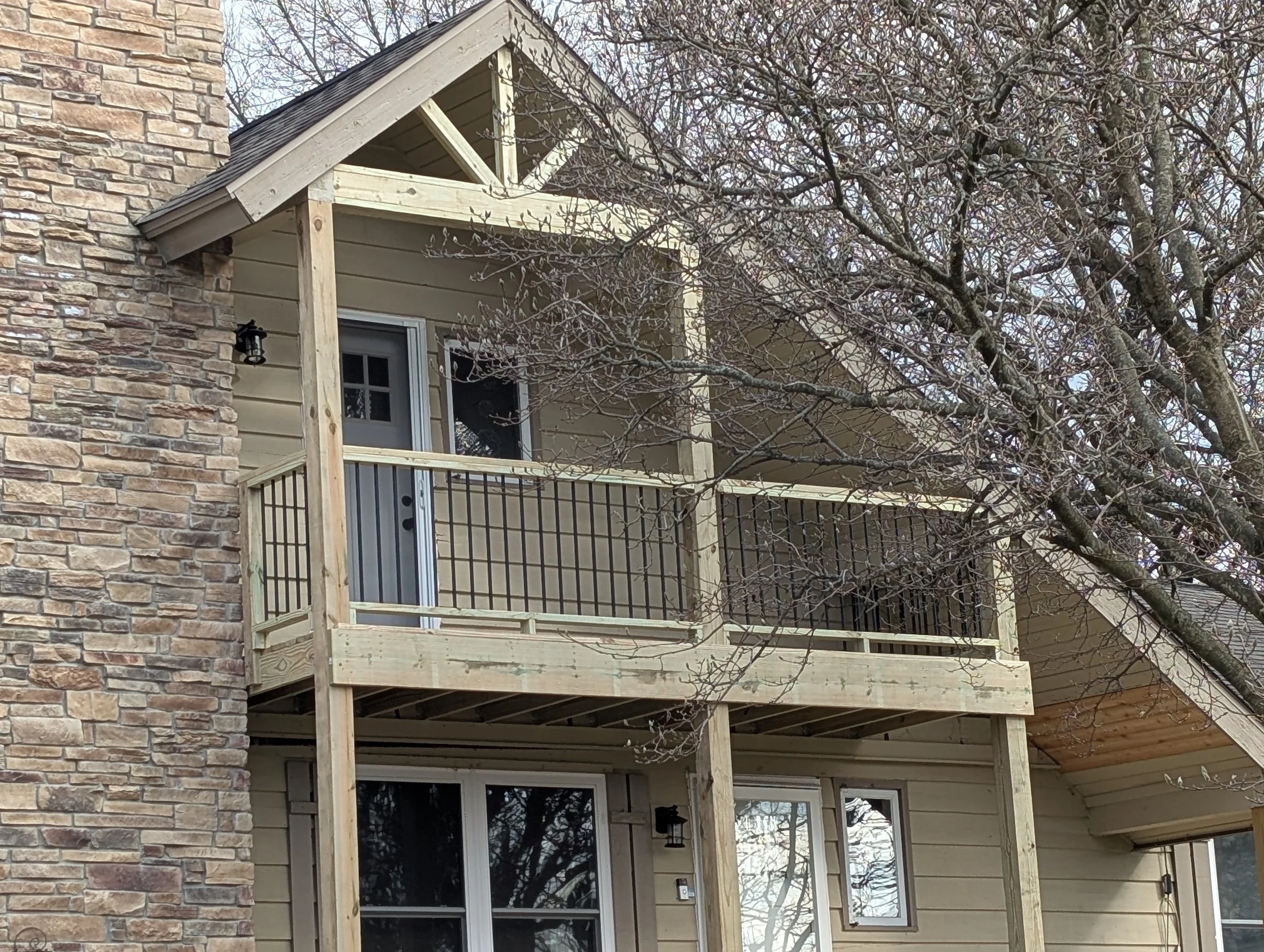 Two-story house with a balcony on the upper level under construction, with wooden railings and a door, next to a stone wall on the left, and a tree with budding branches in front.