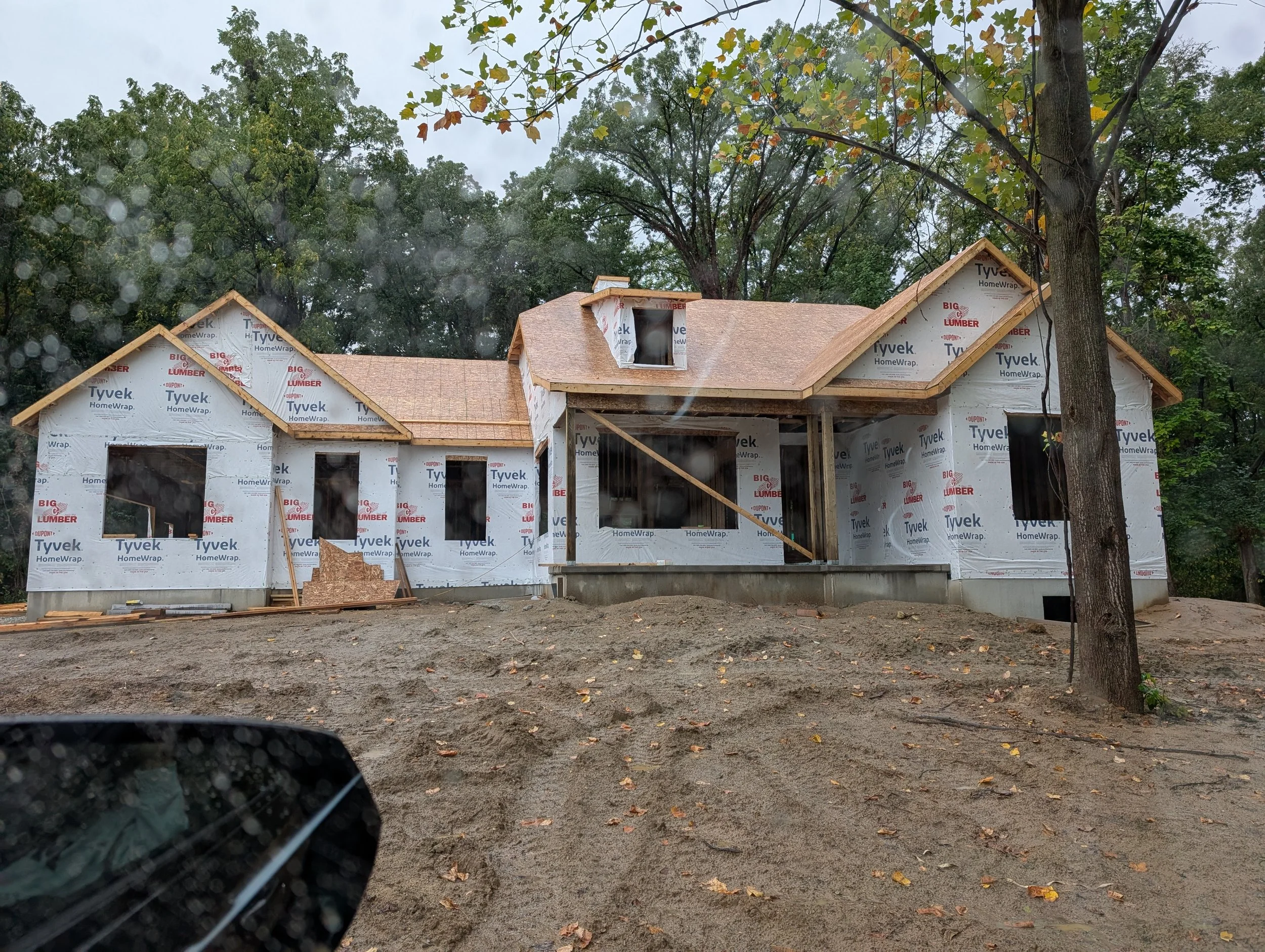 A house under construction wrapped in Tyvek HomeWrap, with a sloped roof and multiple window openings, surrounded by trees and a muddy yard.