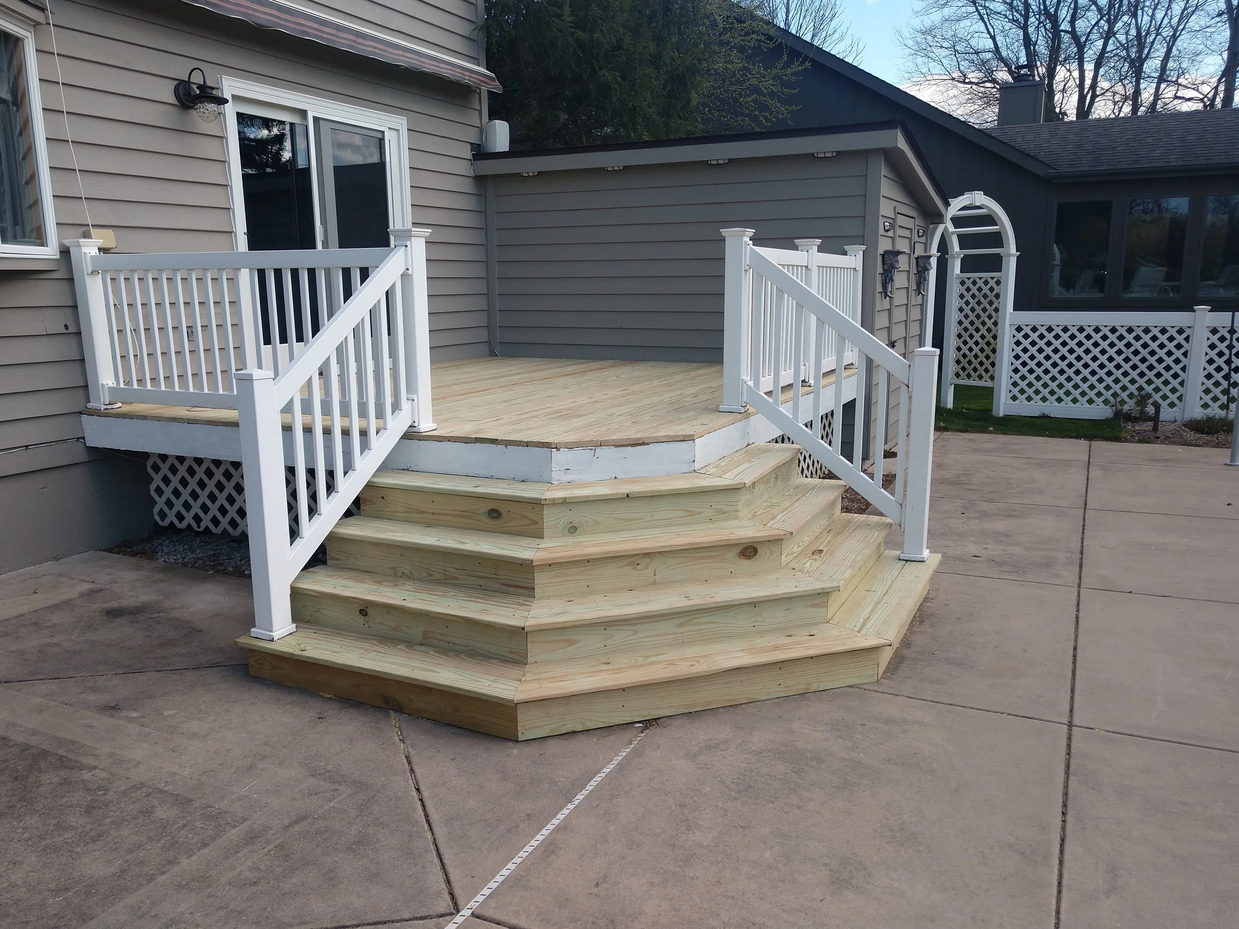 New wooden deck with stairs and white railing in backyard, attached to gray house with sliding glass door.