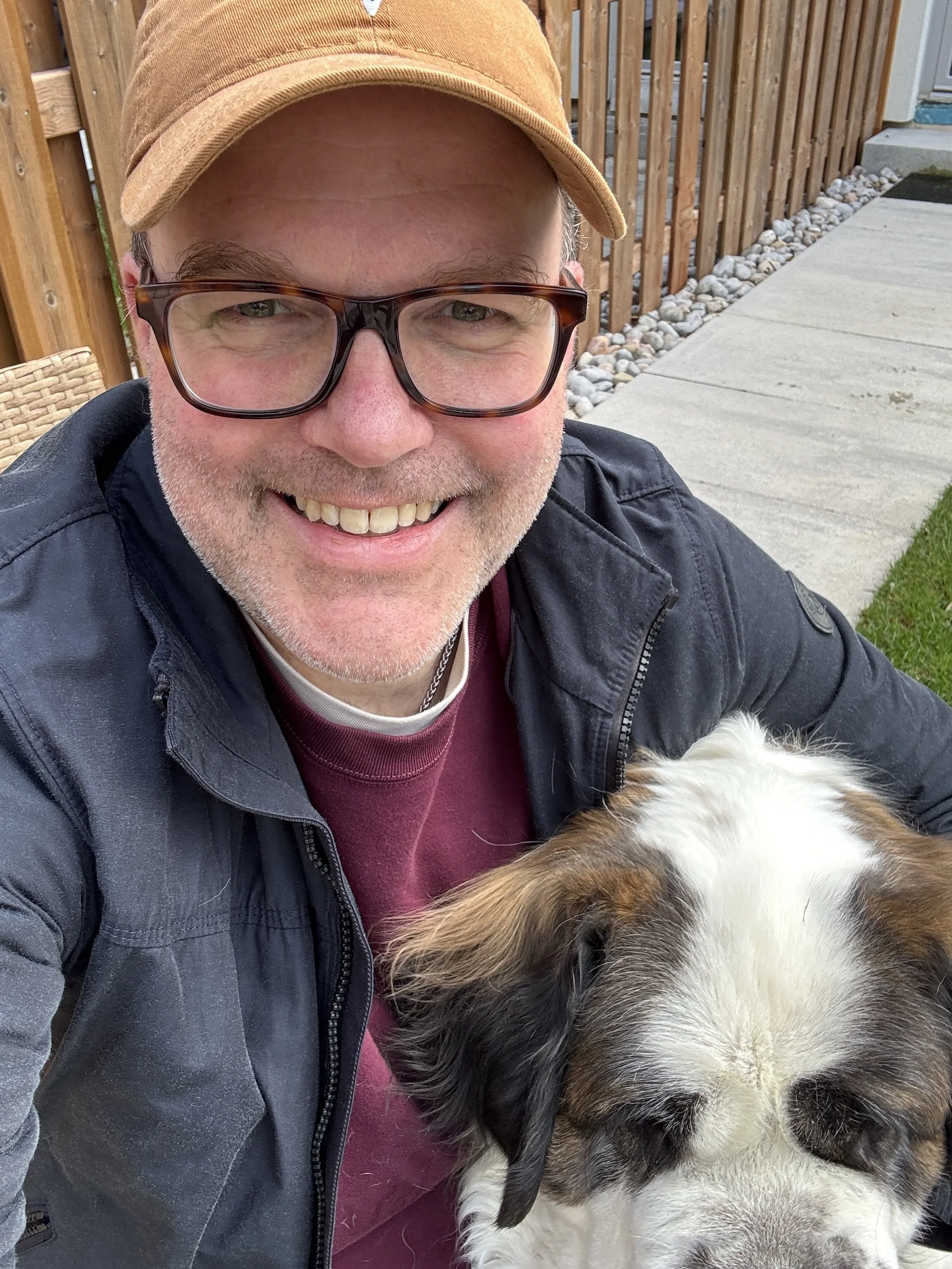 A smiling man wearing glasses, a brown cap, and a black jacket, sitting outside next to a large, fluffy dog with white and brown fur.