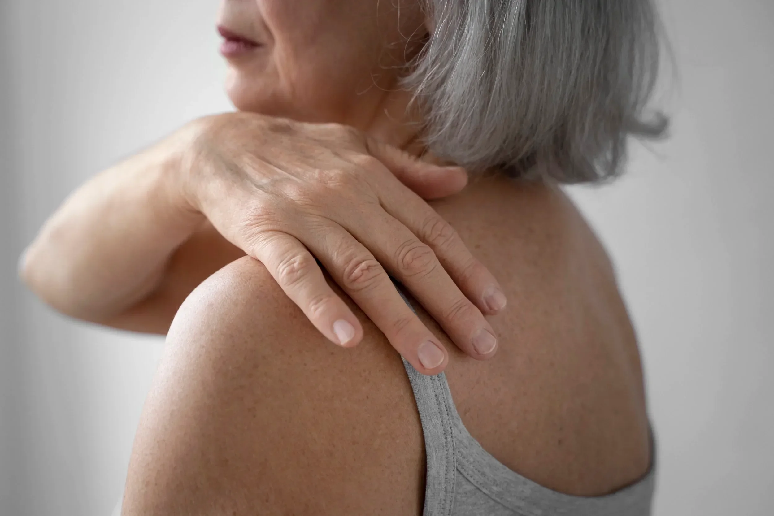 A close-up of a woman with gray hair, wearing a gray tank top, resting her hand on her shoulder. The background is plain and neutral.