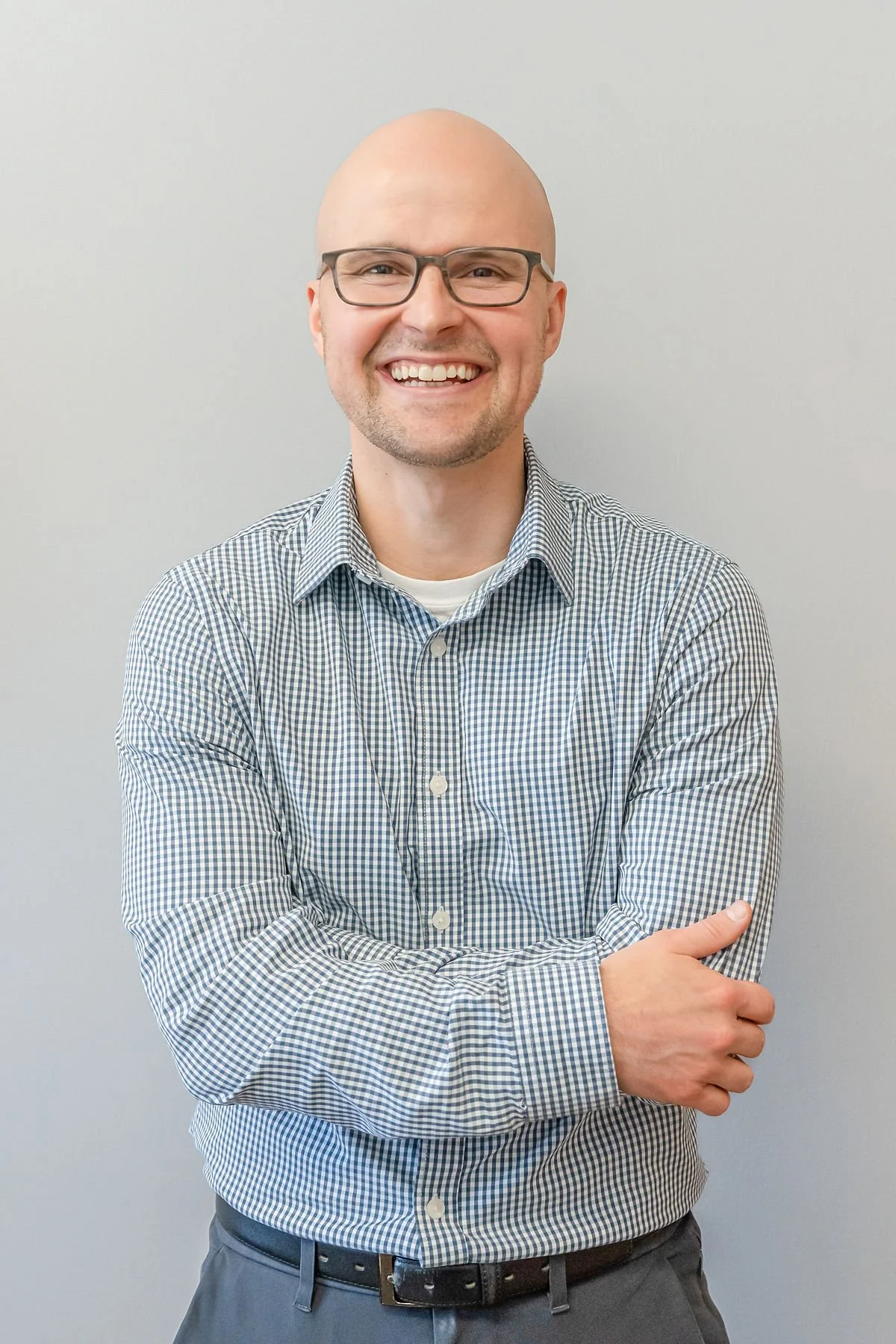 Doctor Eric with glasses, smiling, with a bald head, wearing a checkered shirt, standing against a plain light gray background.