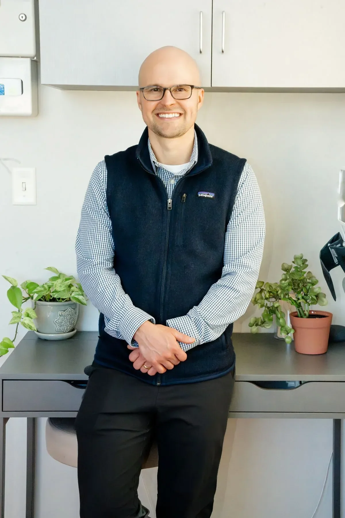 Doctor Eric with glasses, wearing a checkered shirt and a navy vest, smiling and standing in an office with plants on a desk behind him.