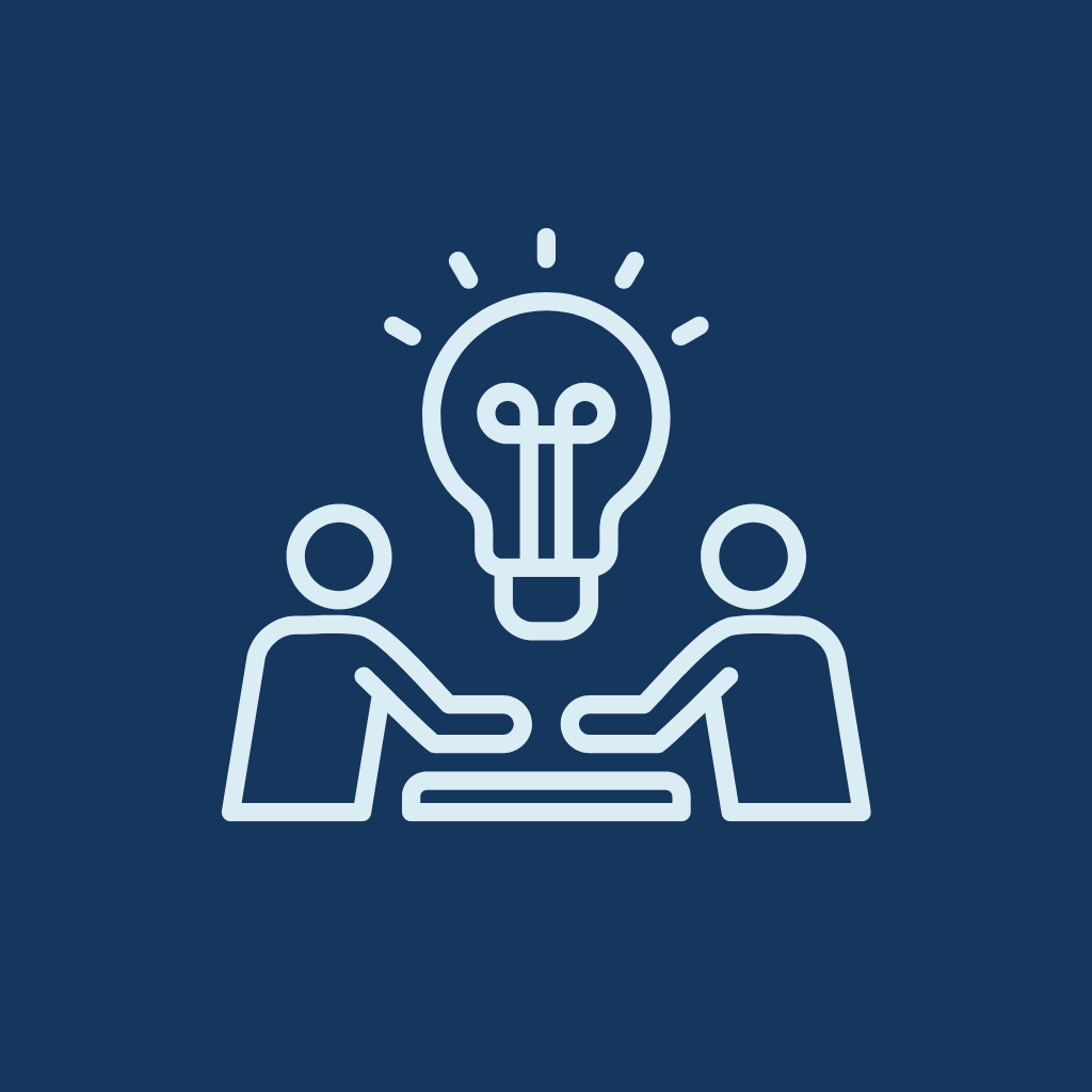 Two people sitting at a table with a large lightbulb above them, symbolizing an idea or discussion.