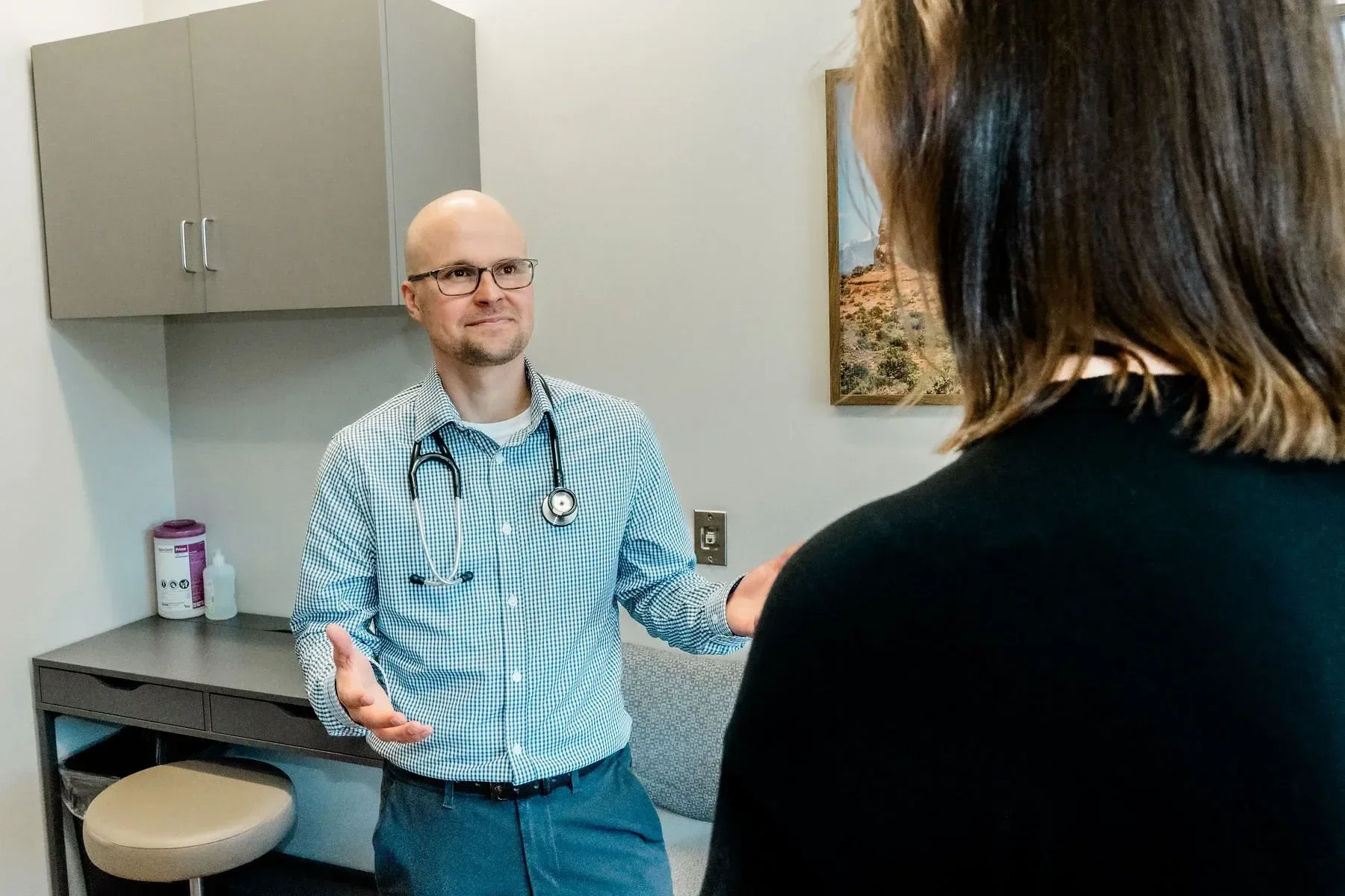Doctor Eric, a male doctor with glasses and a stethoscope around his neck talking to a woman, in a medical office with cabinets and a framed picture of a canyon landscape on the wall.