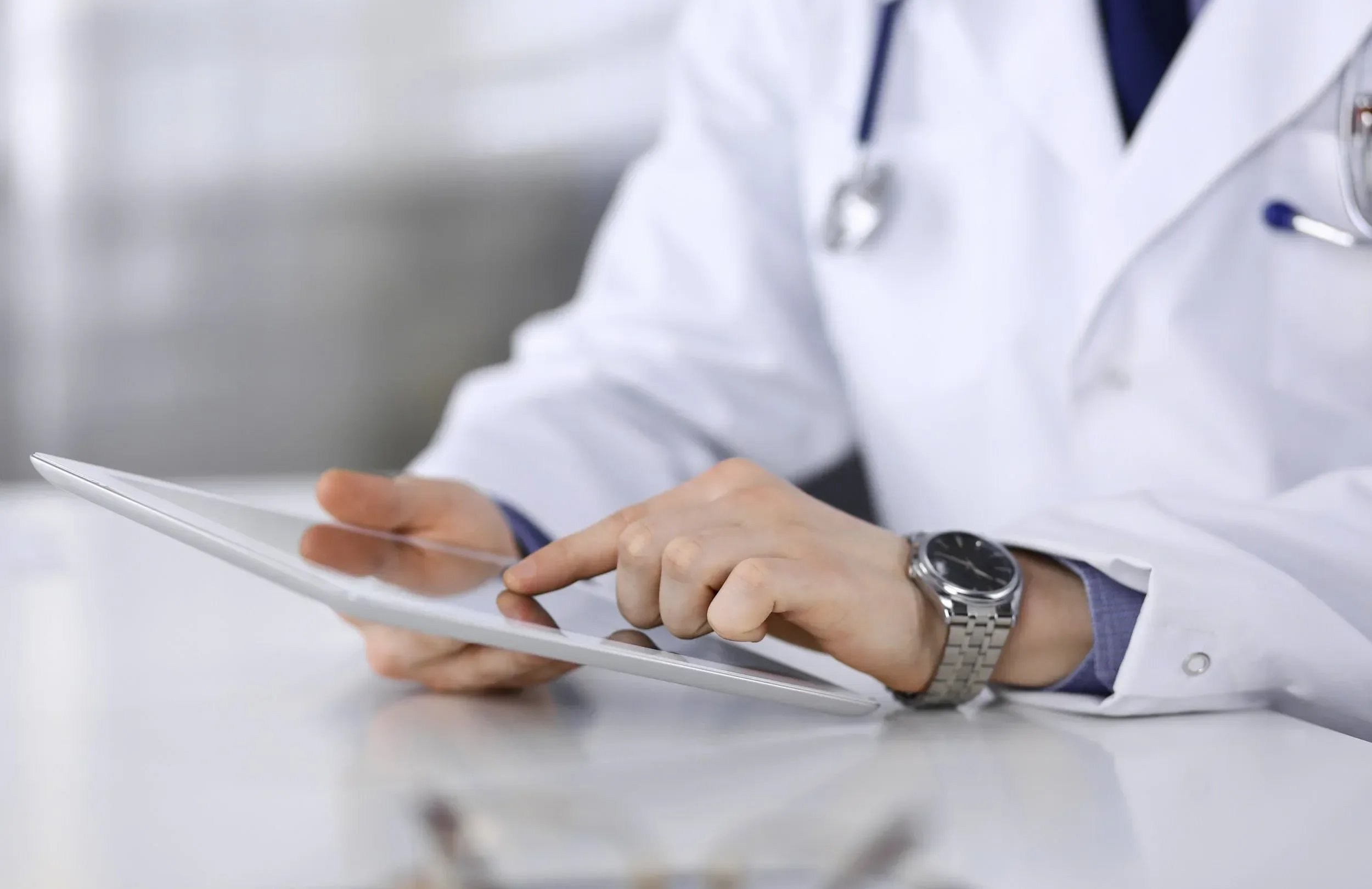 A healthcare professional in a white coat is using a tablet. The person is wearing a wristwatch and a stethoscope hangs around their neck.