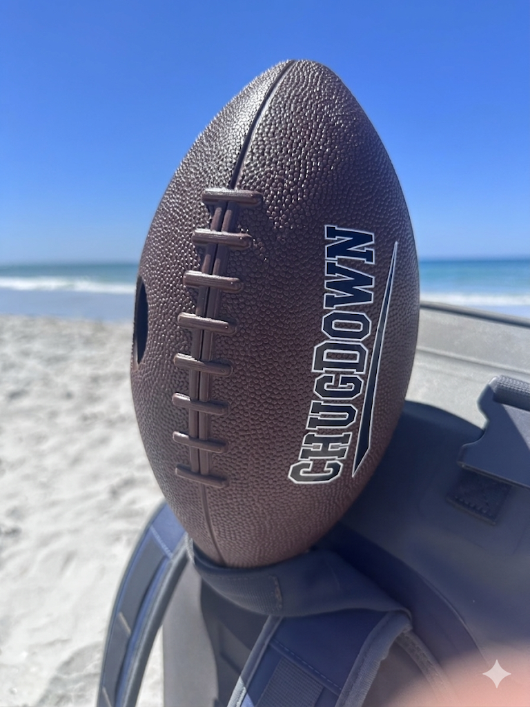 A brown football with the brand name 'CHUGGOV' resting on a beach bag on a sandy beach, with the ocean and blue sky in the background.