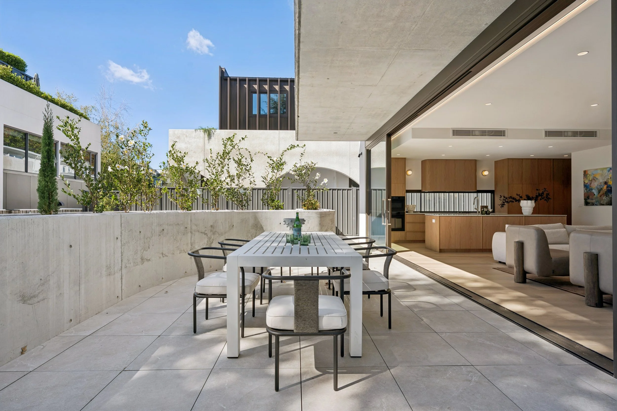 Modern outdoor patio with a white dining table, black chairs, and a glass bottle, adjacent to an indoor living space with wooden cabinets and a painting, set against a blue sky with clouds. Architecturally designed home in Forrest by Thursday