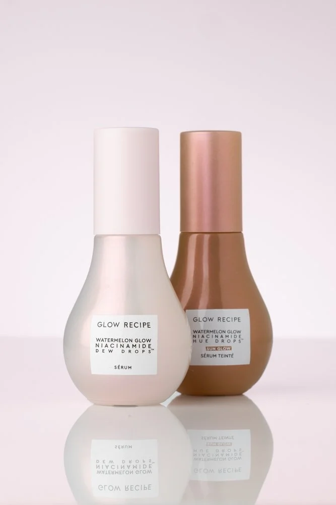 Two bottles of Glow Recipe Watermelon Glow Niacinamide Dew Drops skincare products on a reflective surface against a pink gradient background. product photography