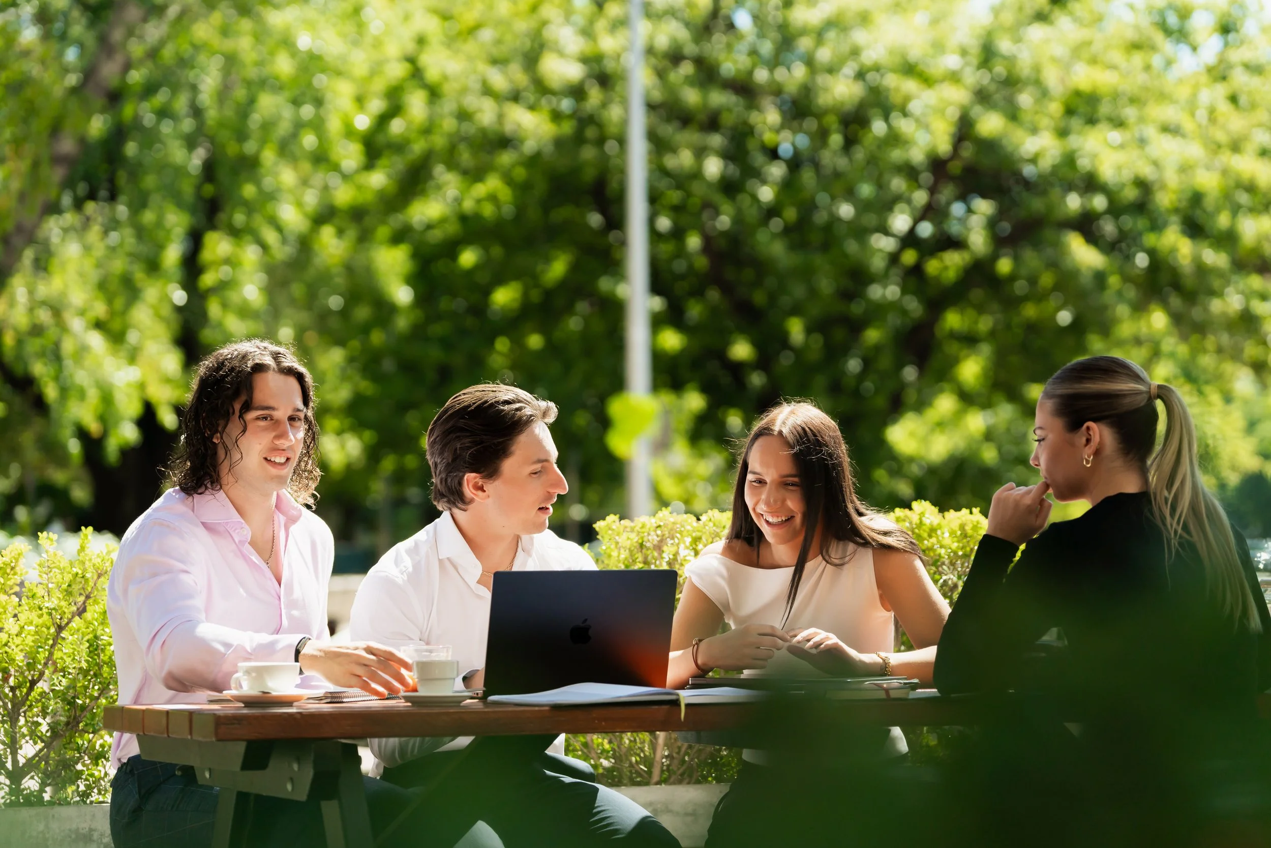 Group of five young adults sitting at an outdoor table, engaged in conversation, surrounded by green trees and bushes on a sunny day.