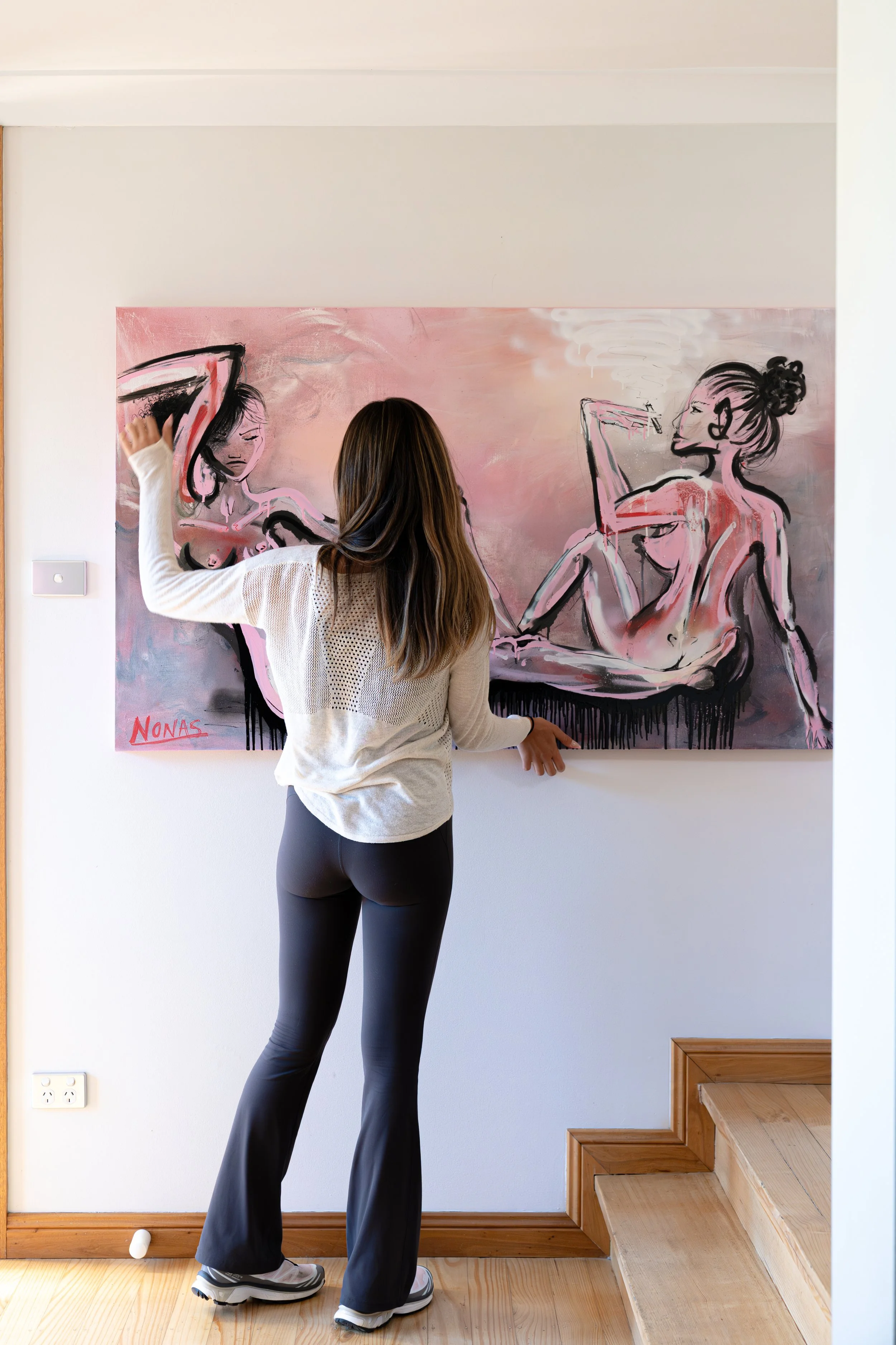 A woman with long brown hair wearing a white long sleeve shirt, black athletic pants, and sneakers, standing in front of a colorful abstract painting on a white wall. The painting features two female figures in a sketchy, artistic style and is signed