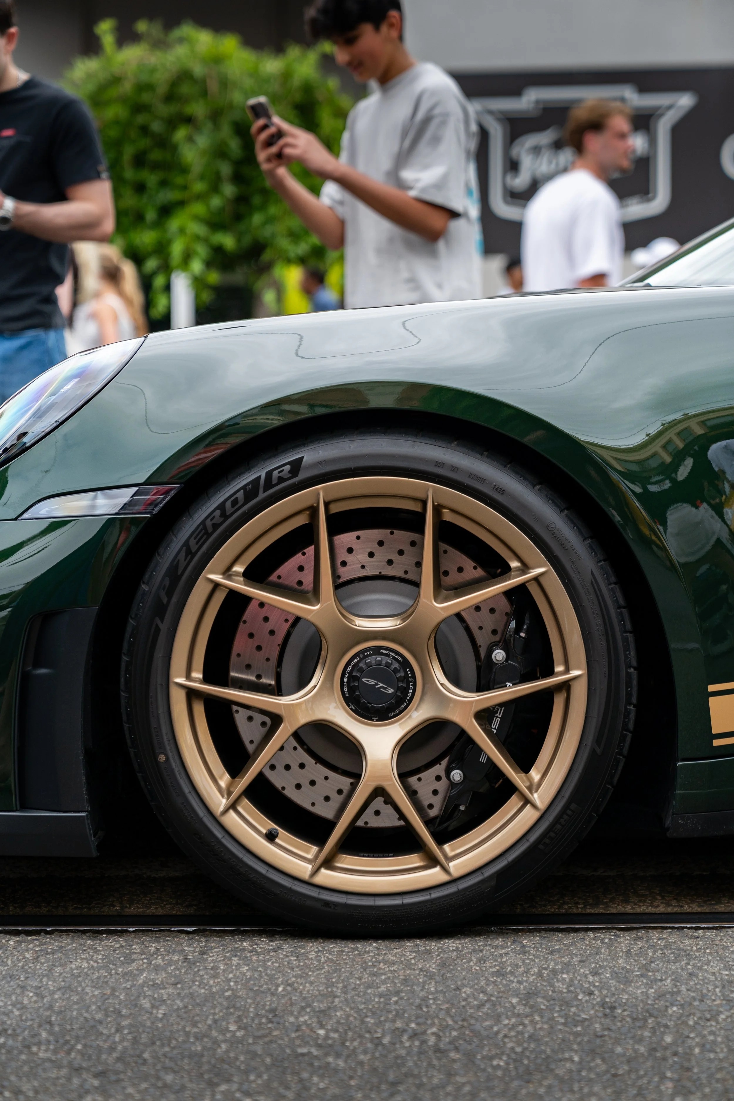 Close-up of a green sports car porsche 911 with gold rims, parked on a street with people in the background at an outdoor event.