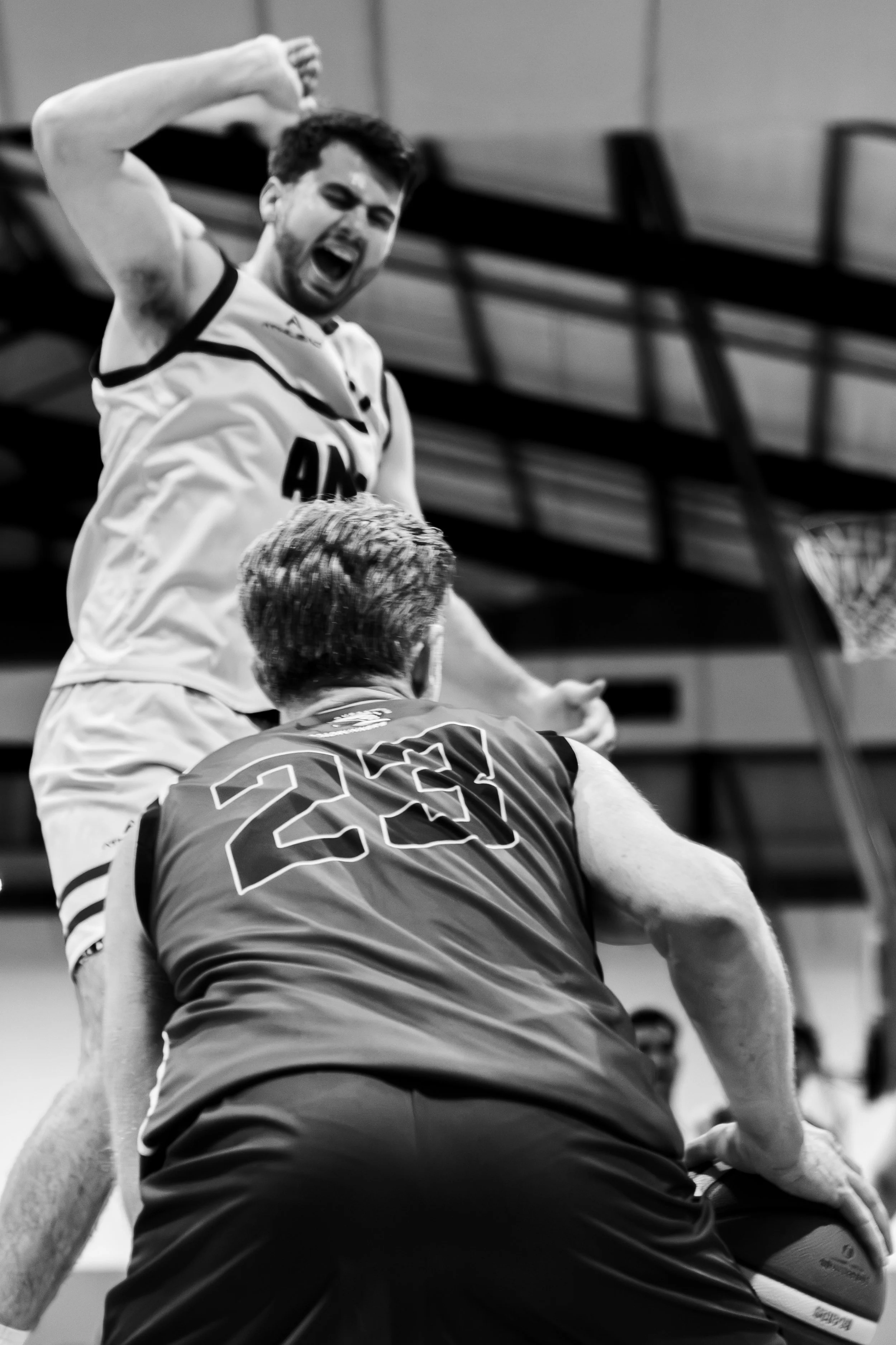 Black and white photo of two male athletes in a basketball match. One athlete is yelling with mouth open, and the other with back facing the camera, wearing a jersey with the number 23. ANU australian national university basketball sports photography