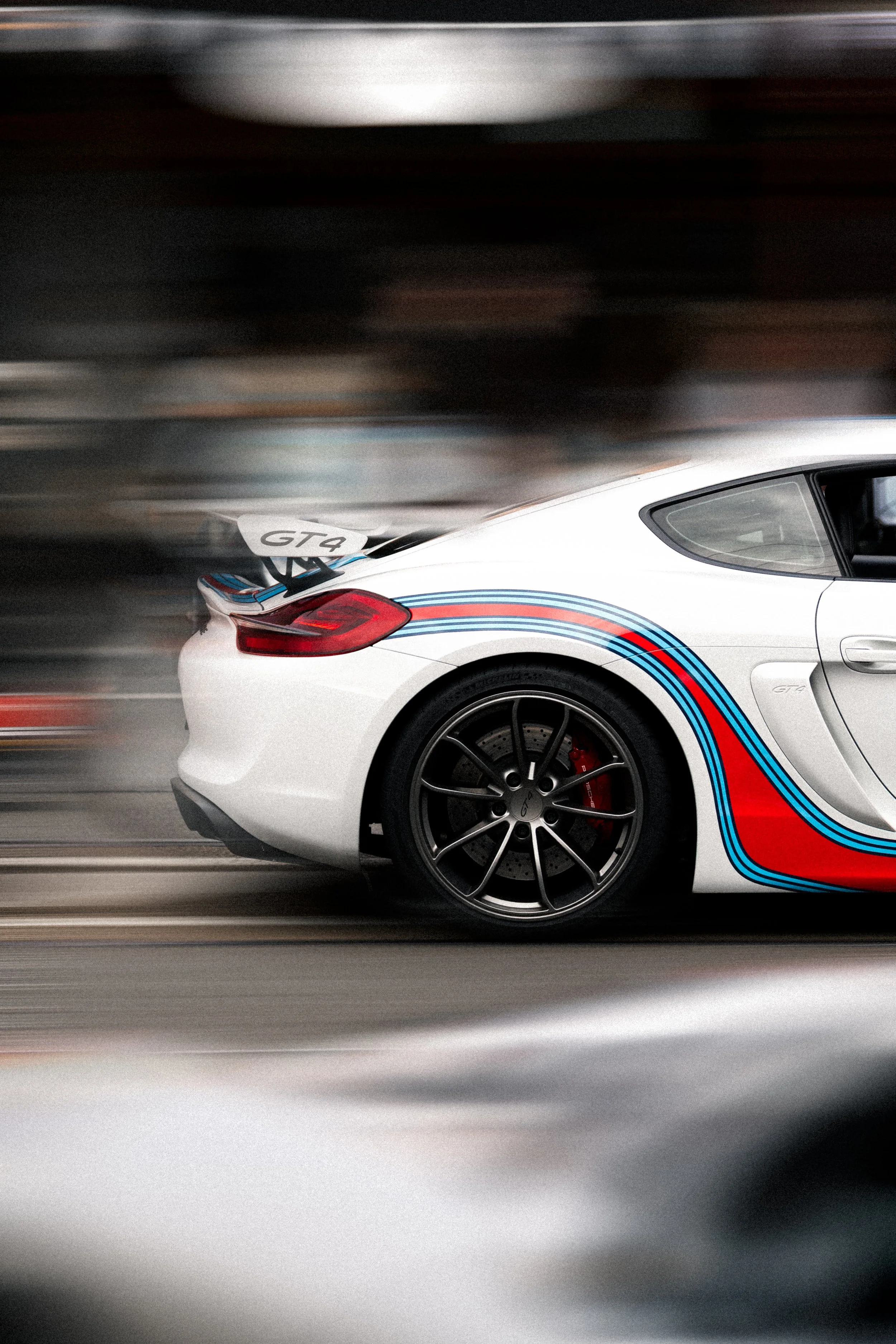 The rear side of a white Porsche GT4 sports car with racing stripes in blue, light blue, and red, in motion on a highway, with a blurred background. melbourne porsche meet motorsport photography