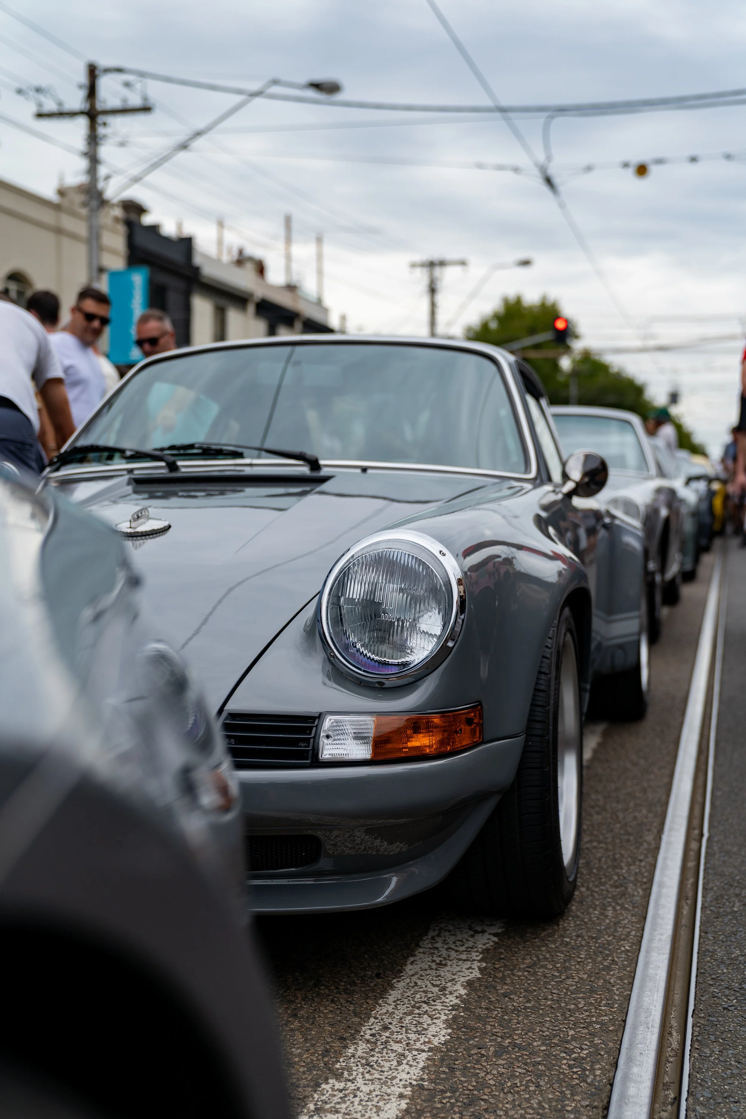 A line of classic silver sports cars porsche 911 930 parked along a street on a cloudy day, with people standing nearby and overhead power lines and traffic lights visible in the background. melbourne australia motorsport photography