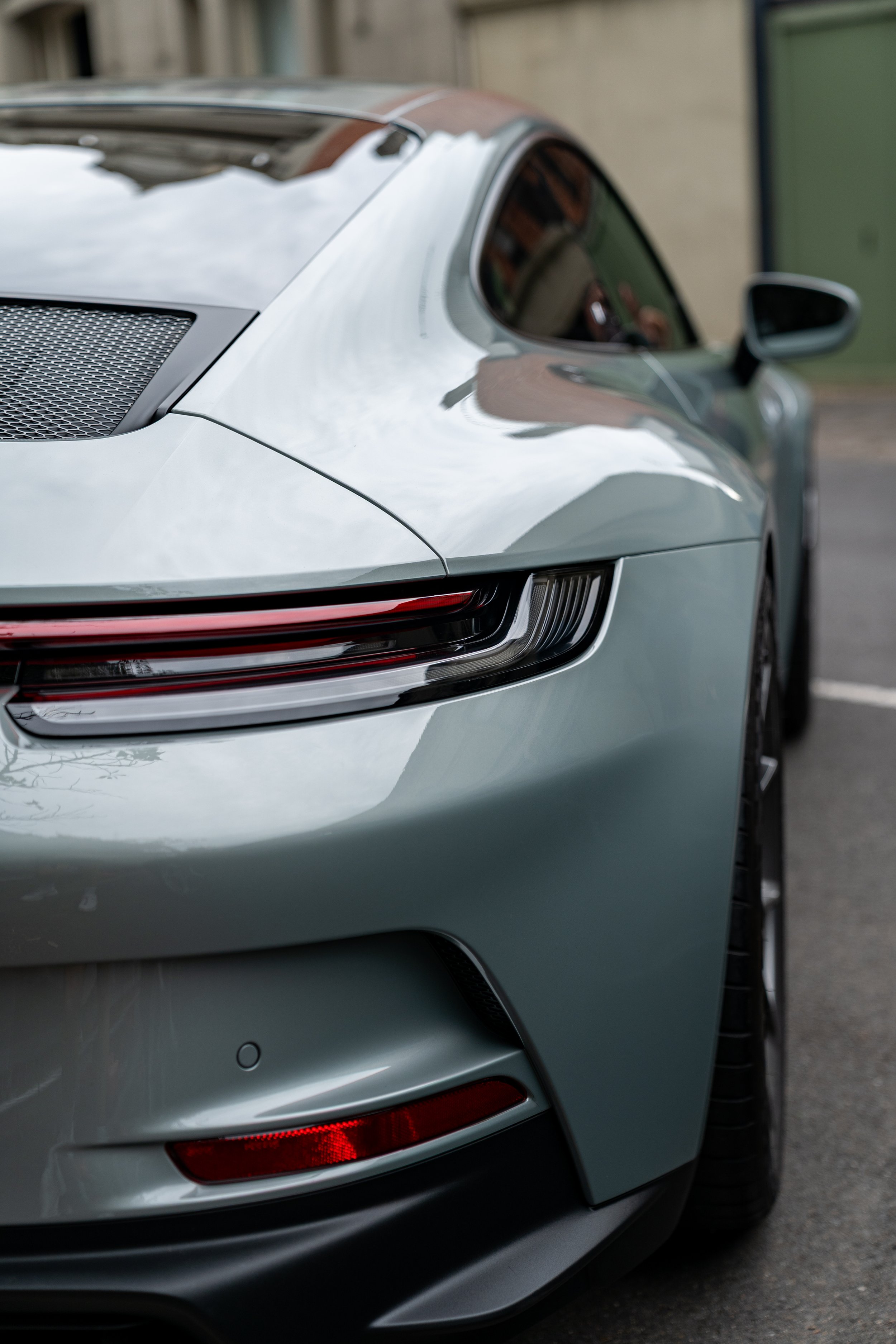 Close-up of the rear and side of a silver sports car posrche 911 GT3 touring parked outdoors, showing the sleek design, taillights, and part of the rear mirror.