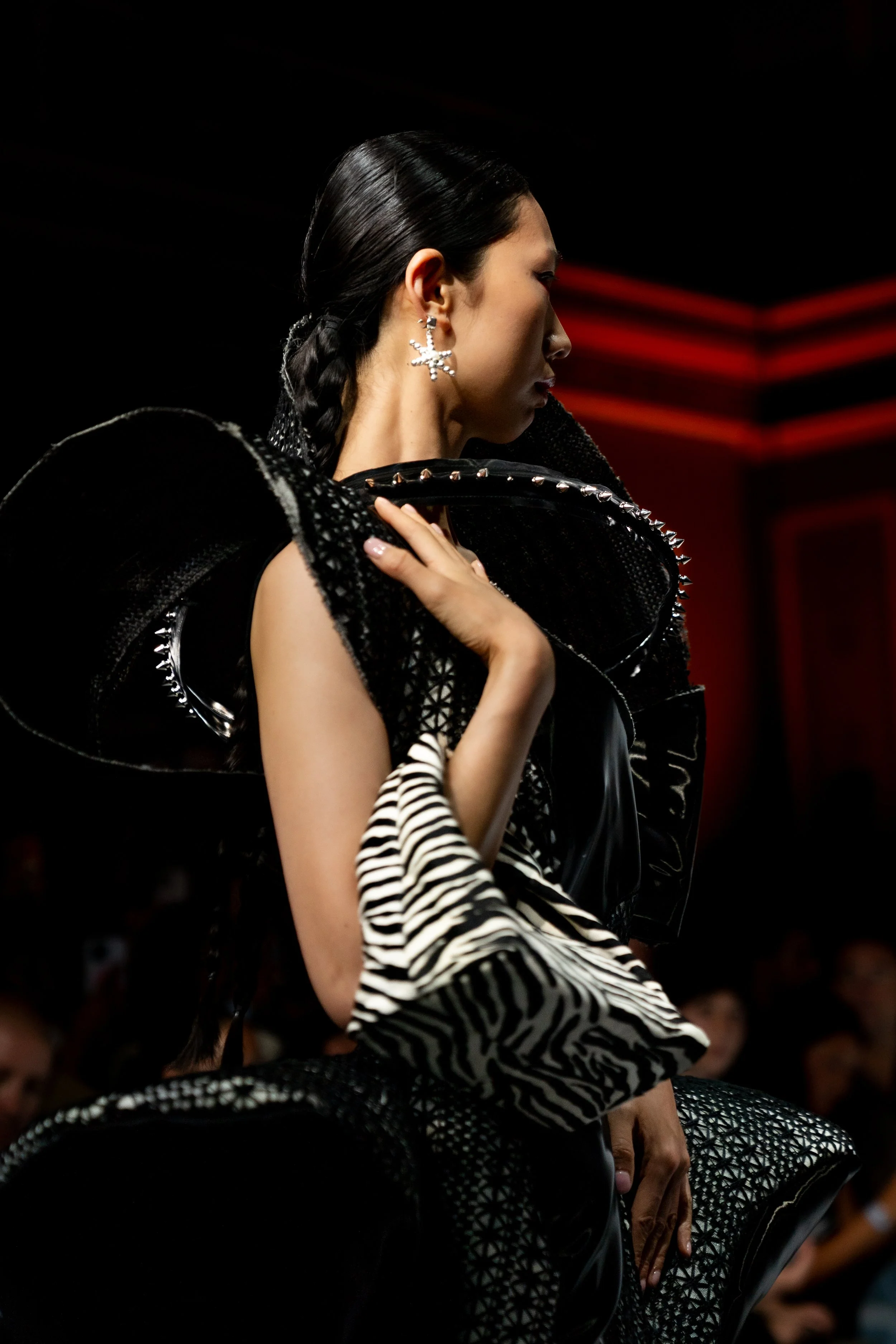 Female model with dark braided hair and star-shaped earrings walking on the runway during a fashion show, wearing a black and white patterned outfit. Baaqiy runway show melbourne fashion festival