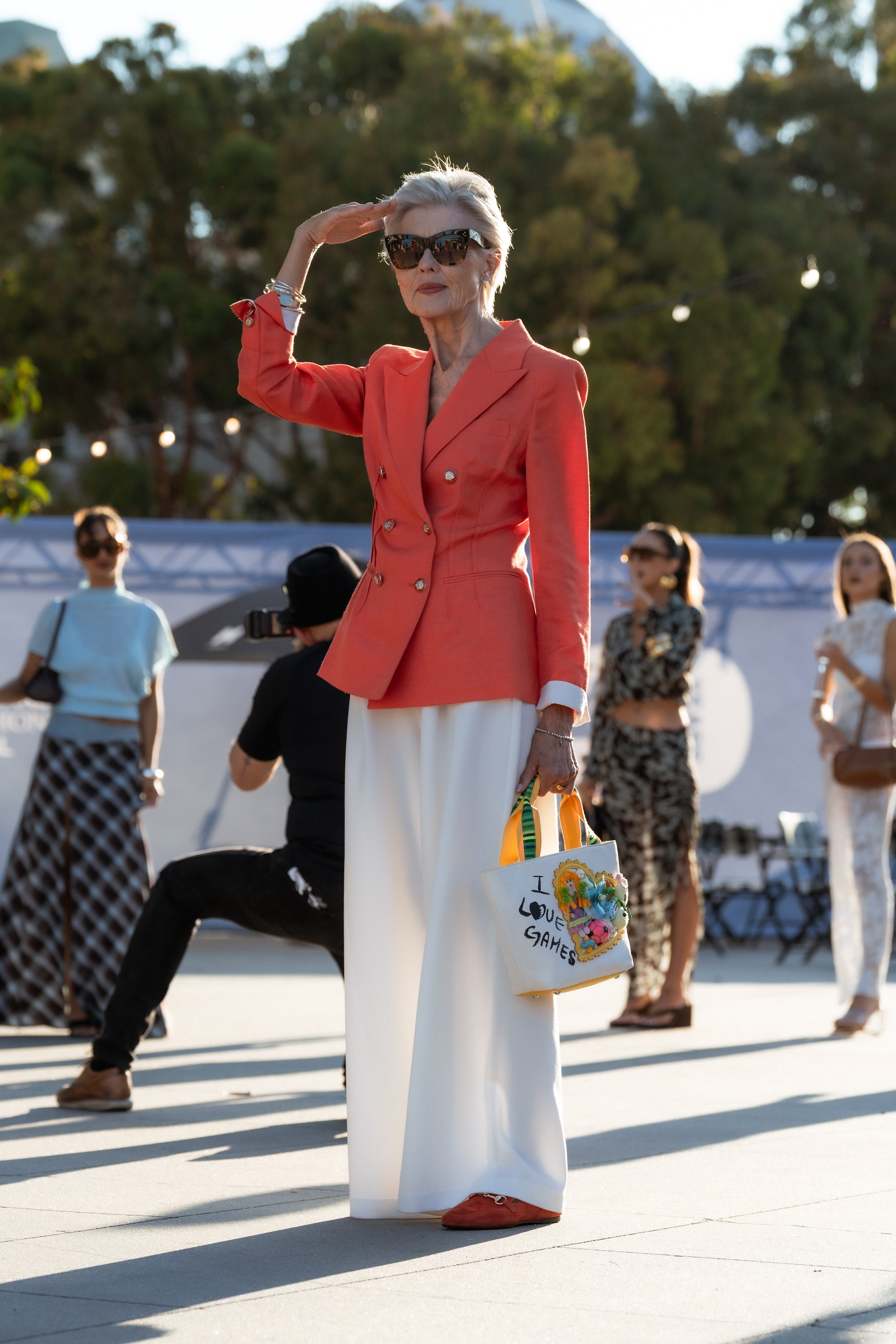 An elderly woman with silver hair, wearing a coral blazer, white wide-leg pants, and sunglasses, salutes with her right hand. She is holding a colorful tote bag decorated with plush toys and the phrase 'I LOVE GAMES' written on it. Several women in t