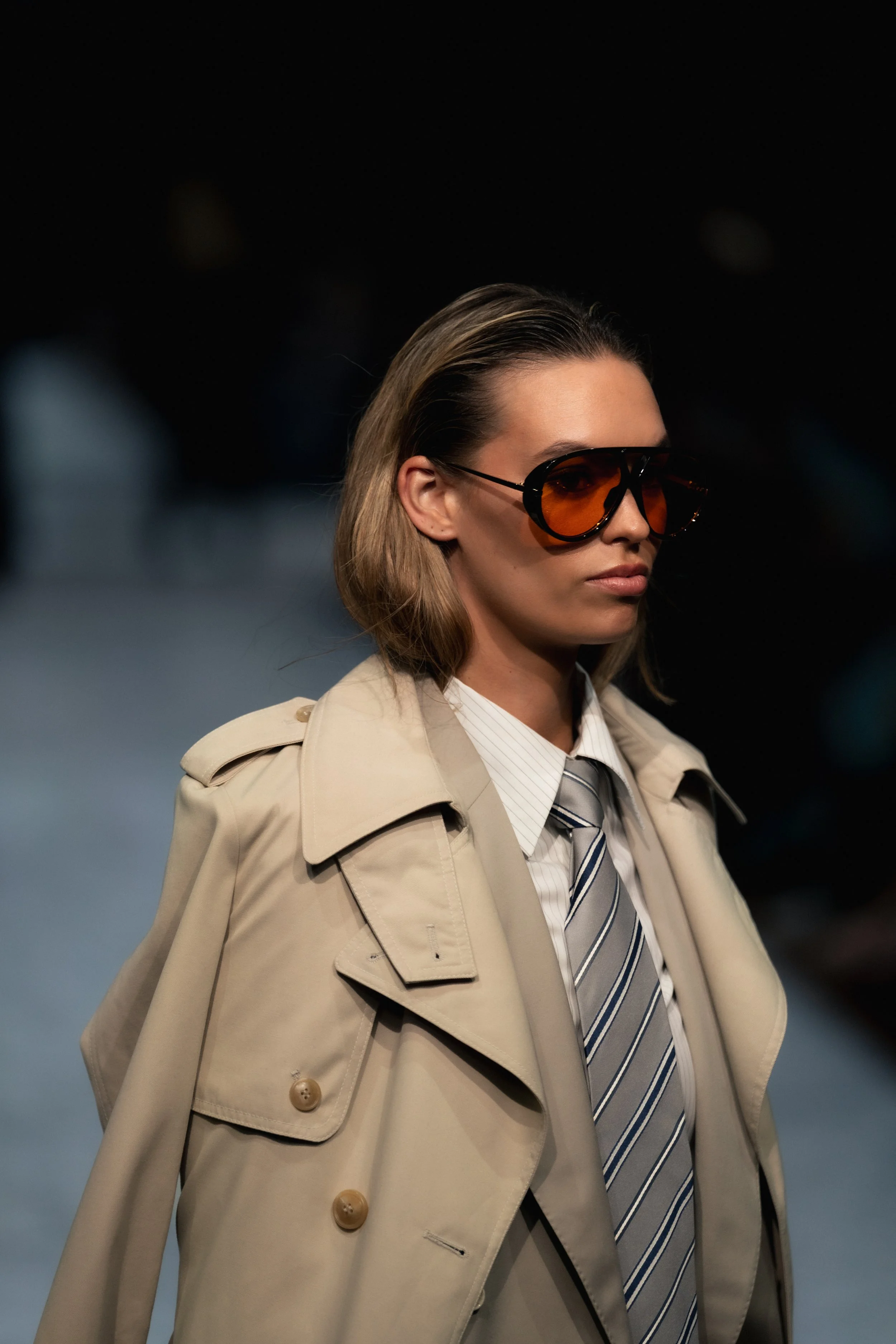 Woman wearing a beige trench coat, white shirt, striped tie, and large sunglasses with orange tinted lenses, standing outdoors at night. melbourne fashion festival forage australia runway show